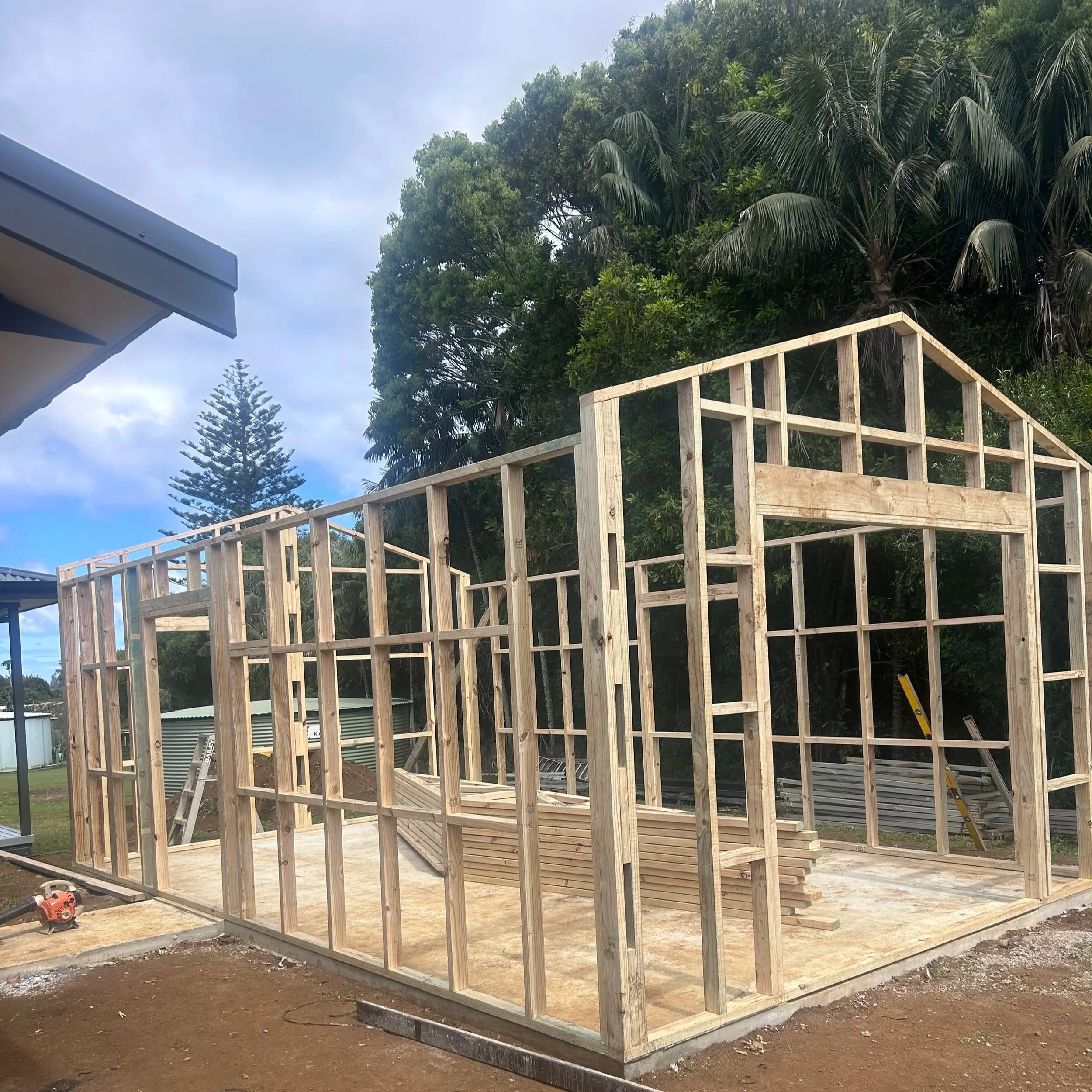 Construction site with a wooden frame structure being built, surrounded by green trees and cloudy sky.