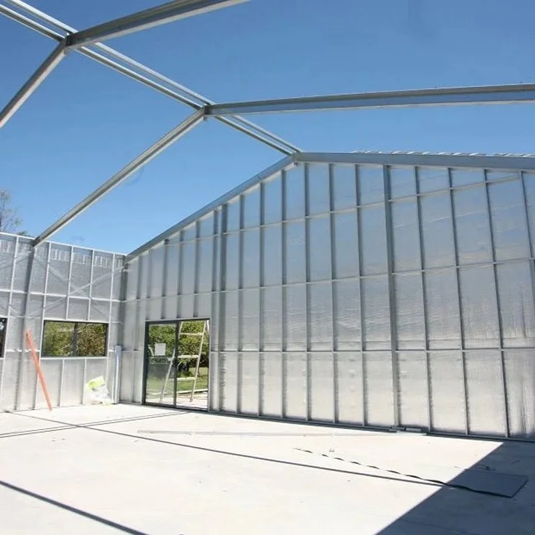 The image shows the interior of a greenhouse under construction with metal framing and transparent panels, sunlight casting shadows on the concrete floor.