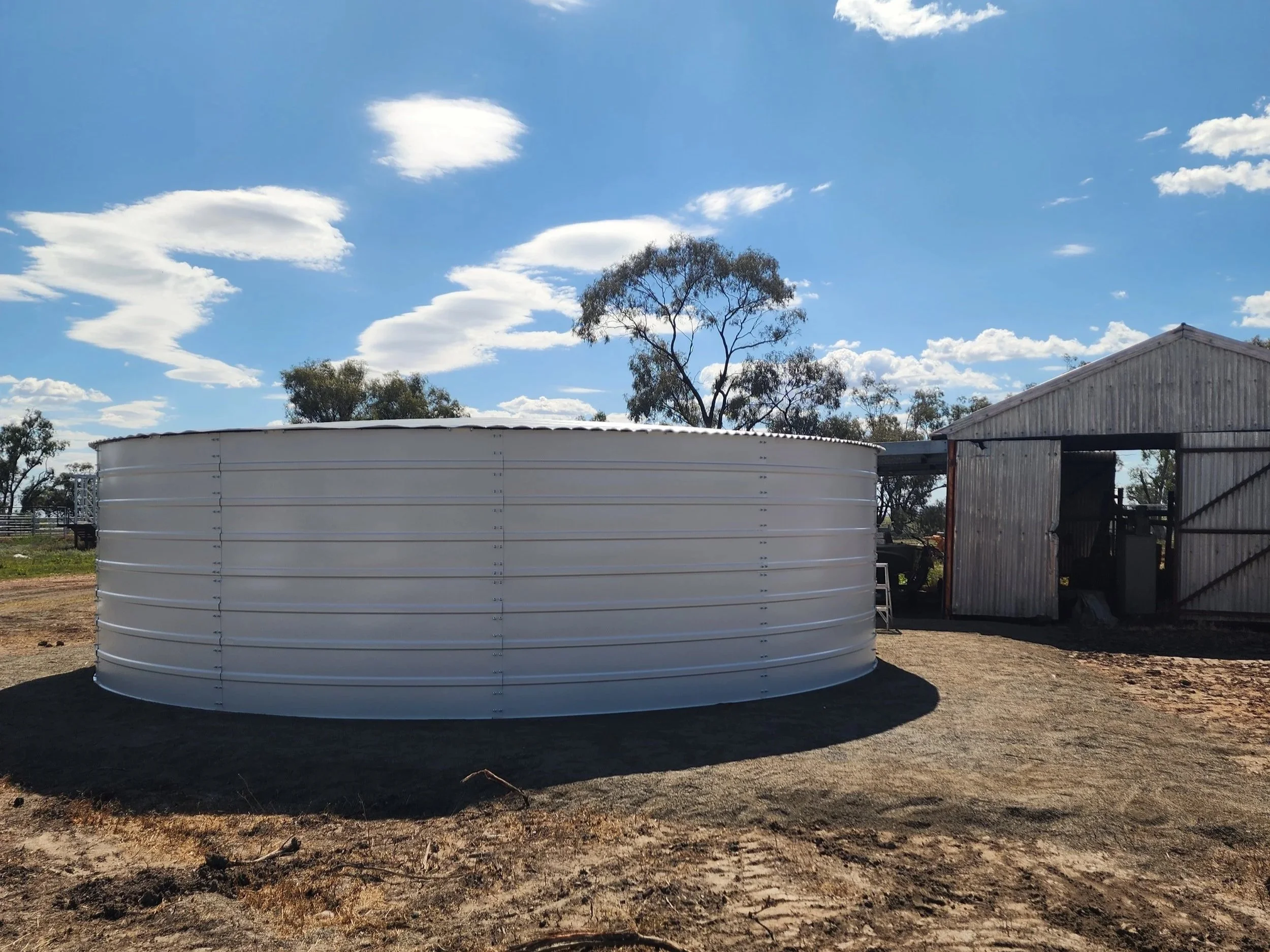 A rural farm scene with a large white water tank, some trees in the background, and an old metal barn building to the right. The sky is partly cloudy with several fluffy clouds.