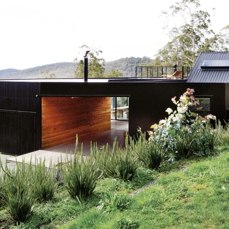 Modern house with black exterior and wooden-covered garage, surrounded by lush greenery and a garden with flowering plants, in a hilly area.