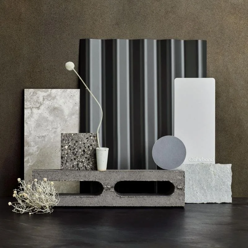 Sample materials and textures for interior design, including concrete blocks, marble slabs, a white ceramic vase with a flexible stem, paint swatches, and a piece of corrugated metal.