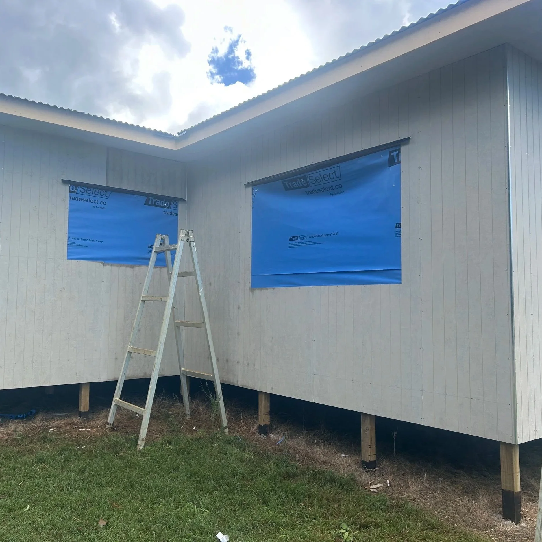 Construction site with a partially built house, blue house wrap on the windows, wooden stilts elevating the house, and a ladder leaning against the wall. Grass and dirt are visible on the ground, with cloudy sky overhead.