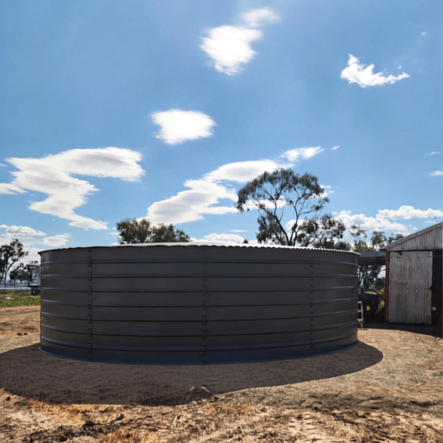 A large, round metal water tank on a dirt ground with a shadow cast below it, set in a rural area with scattered trees, a small shed, and a partly cloudy sky.