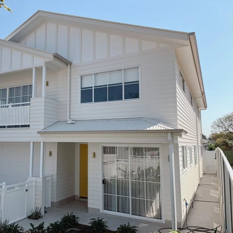 A white two-story house with a yellow front door, large front window, small balcony, and a fenced yard.