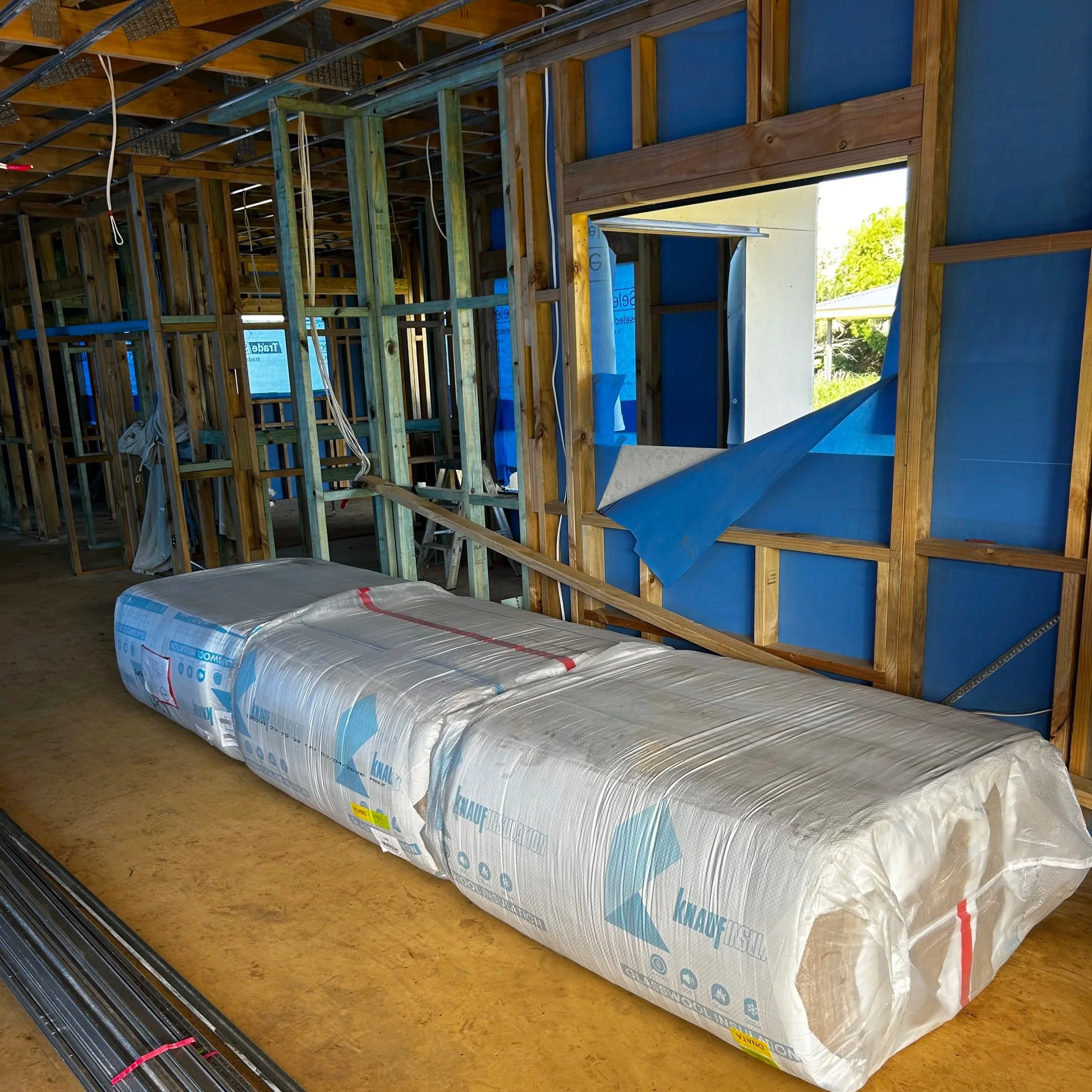 Large rolls of insulation material in clear plastic packaging stacked on a wooden floor inside a partially constructed building.