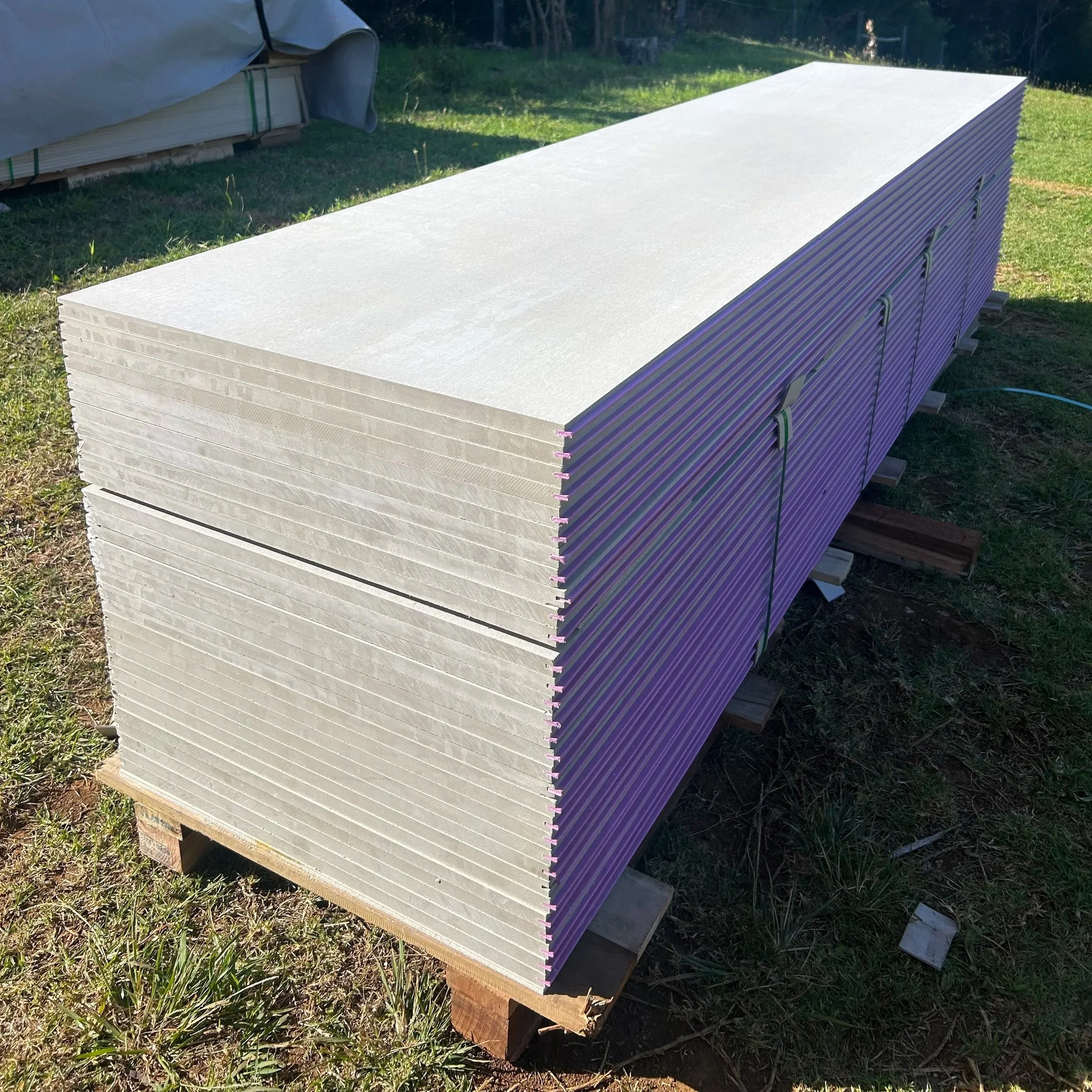 Stack of drywall sheets outdoors on grass with trees in the background.