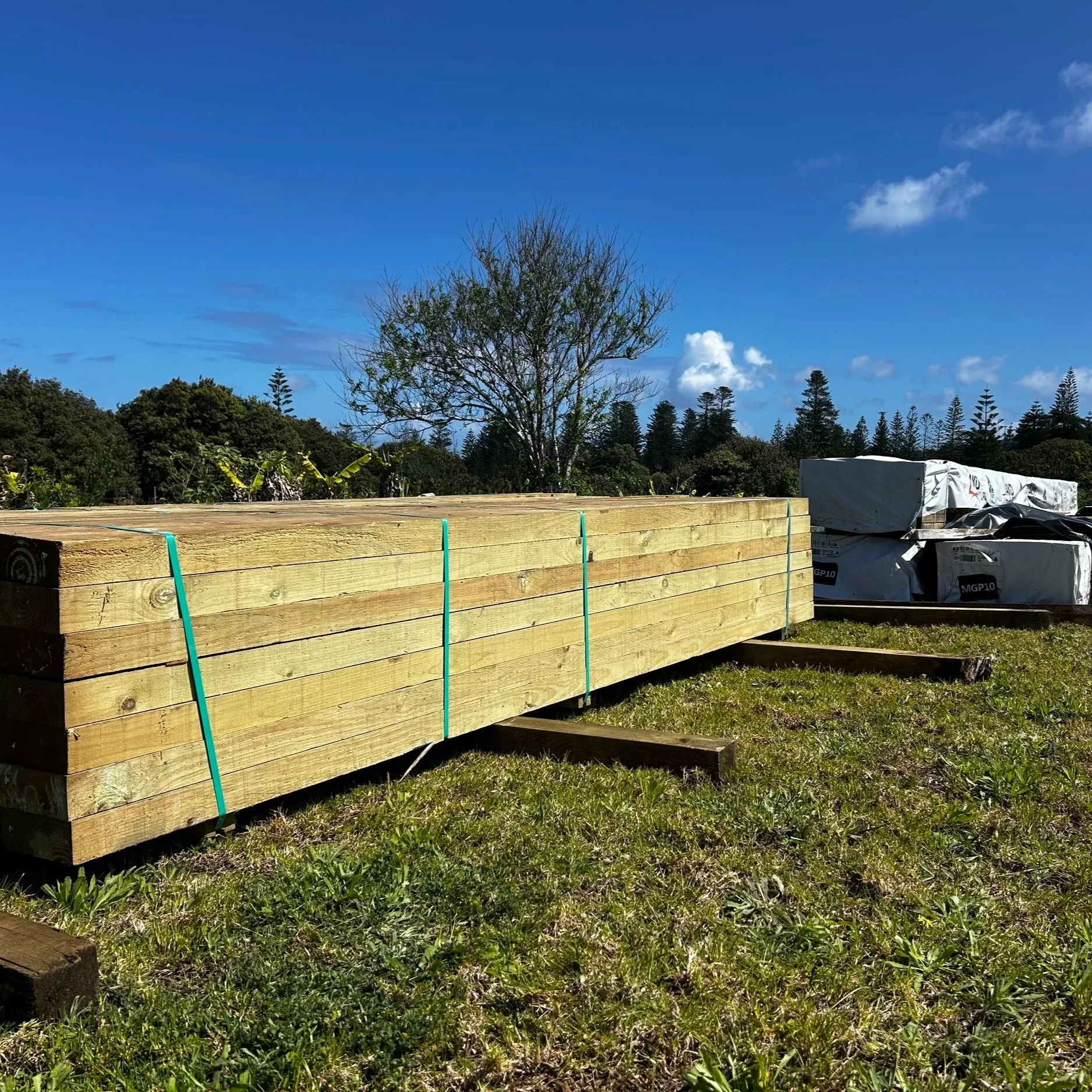 Stacked wooden planks outdoors with construction materials and trees in the background under a blue sky with a few clouds.