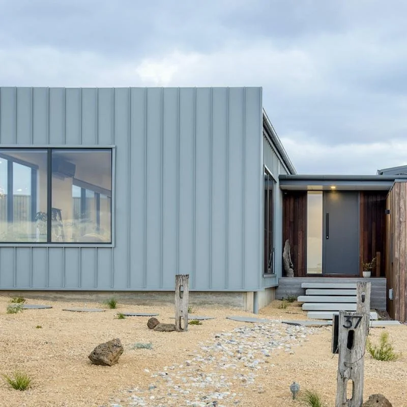 Modern house with metal siding, large windows, and a wooden entryway, situated in a sandy landscape with rocks and sparse vegetation.