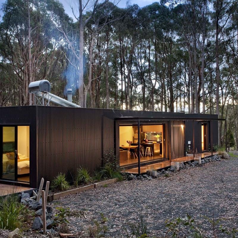 A modern, black metal house with large sliding glass doors, surrounded by trees, with warm interior lighting visible from outside during dusk.