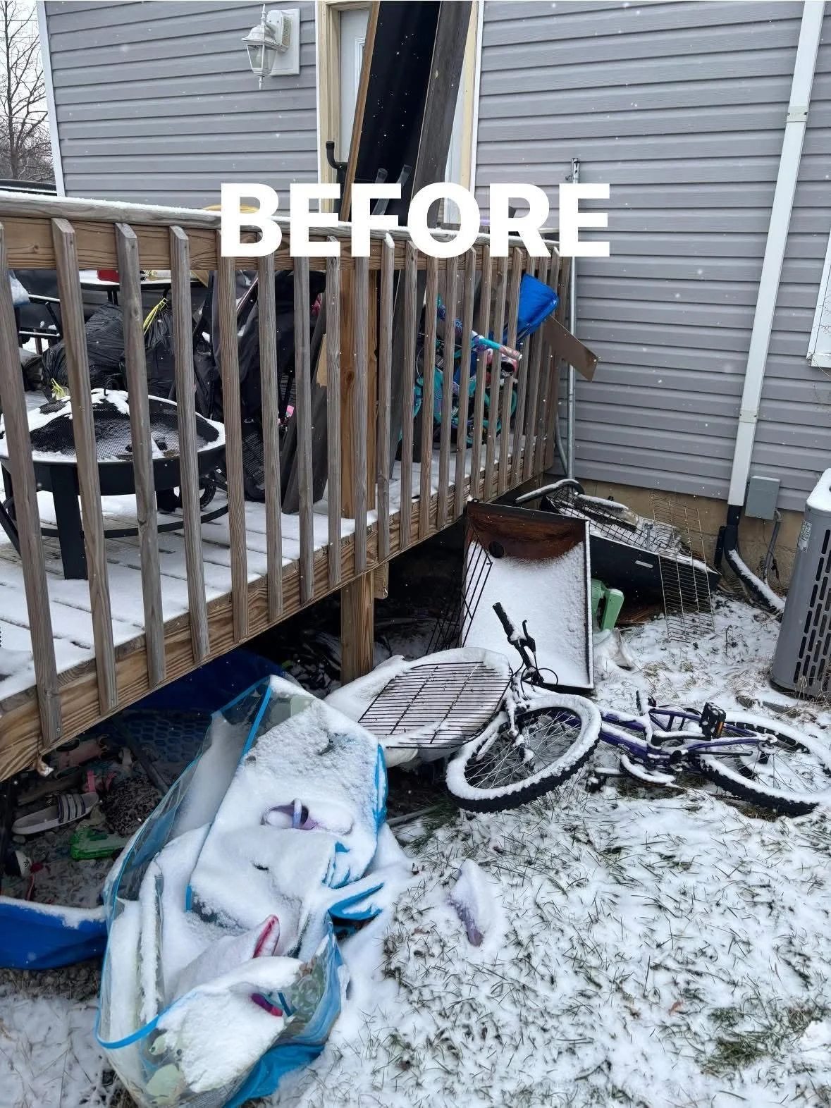 Backyard scene with a small wooden deck covered in snow, cluttered with a bicycle, a grill, a laundry rack, and other outdoor items. Snow is on the ground, and a house with grey siding is visible in the background.