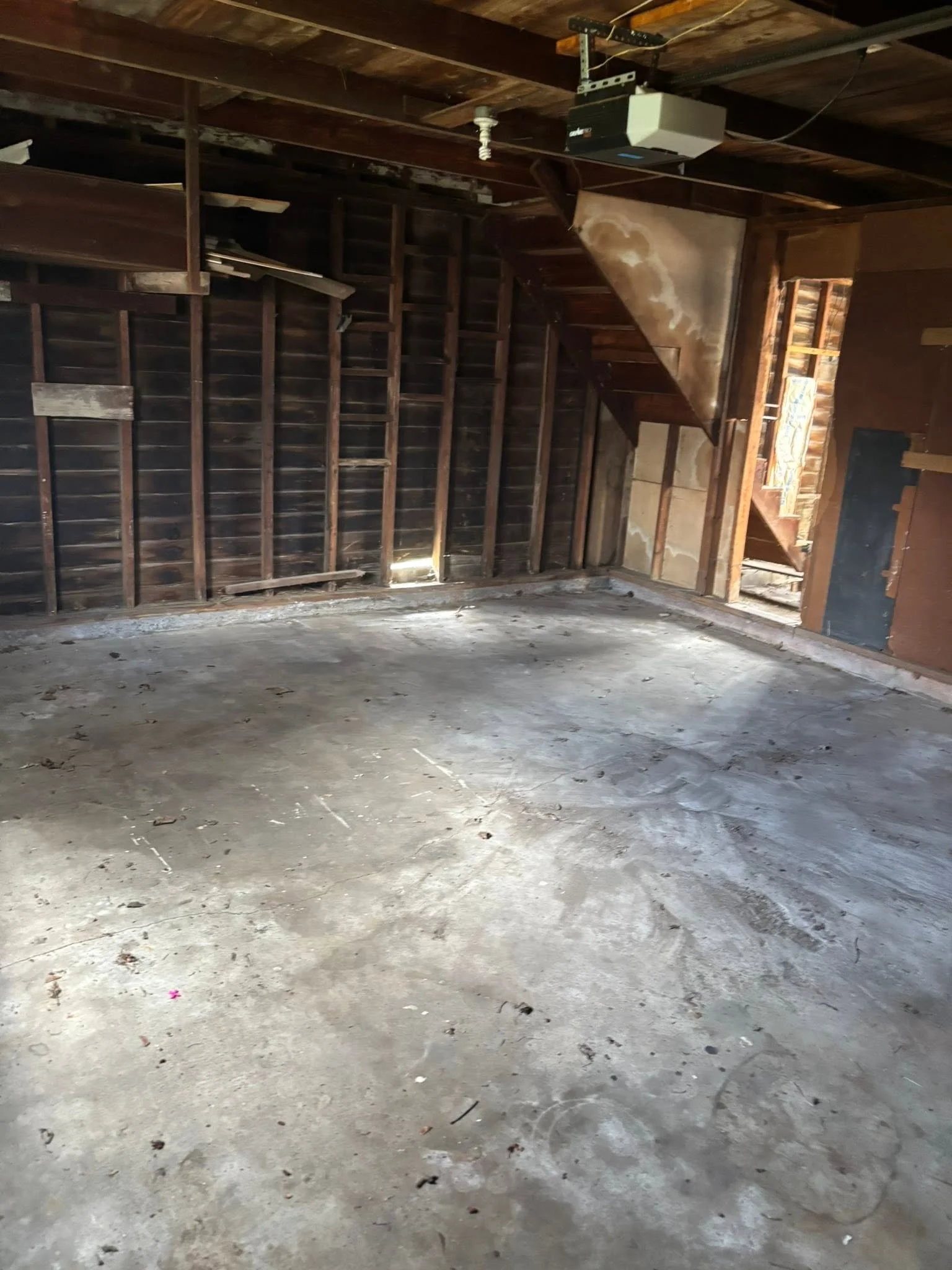 Empty unfinished garage with concrete floor, exposed wooden wall studs, and a garage door opener mounted on the ceiling.