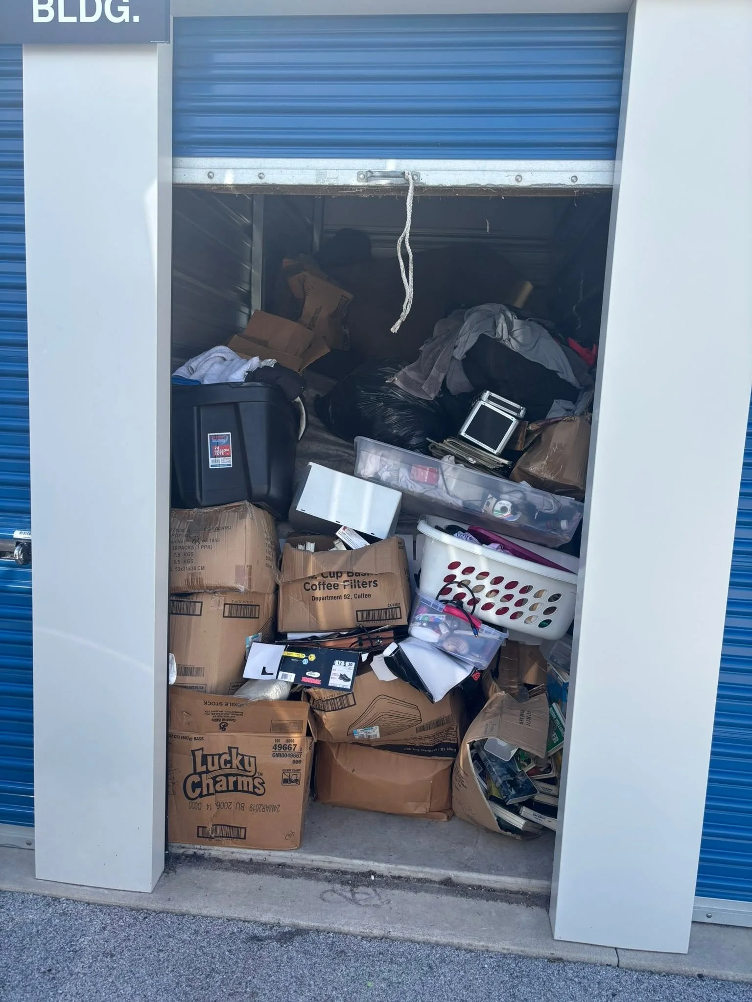 A storage unit with its door partially open, filled with boxes, used items, and household goods, including a Lucky Charms cereal box and various miscellaneous objects.