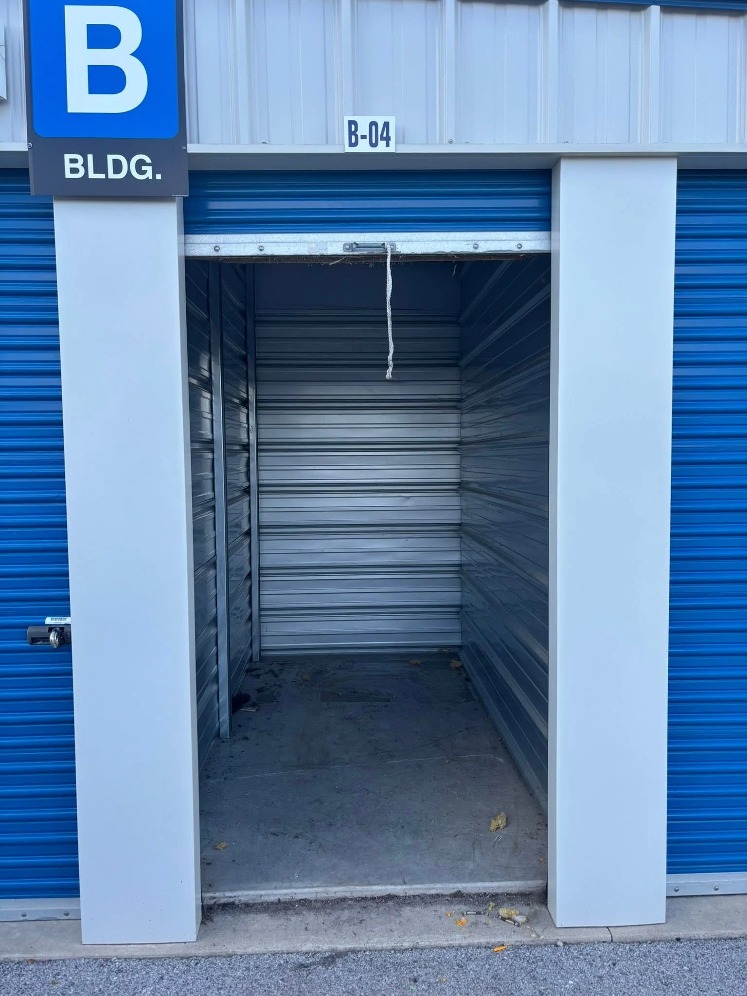Empty storage unit with partially open blue metal roll-up door, white shelving, and a concrete floor, situated on an asphalt surface.
