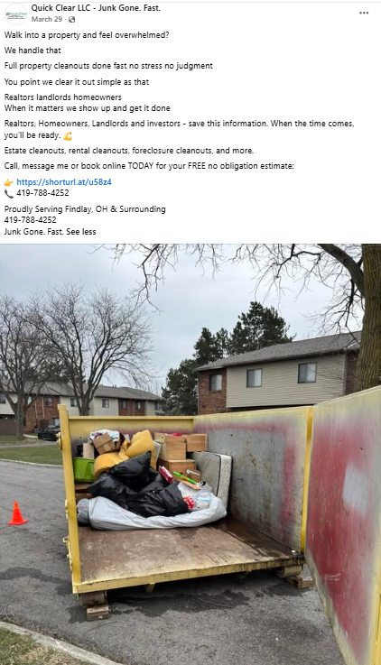 A large yellow dumpster filled with trash, including black garbage bags, cardboard boxes, and miscellaneous debris, situated on a residential street with trees and houses in the background.