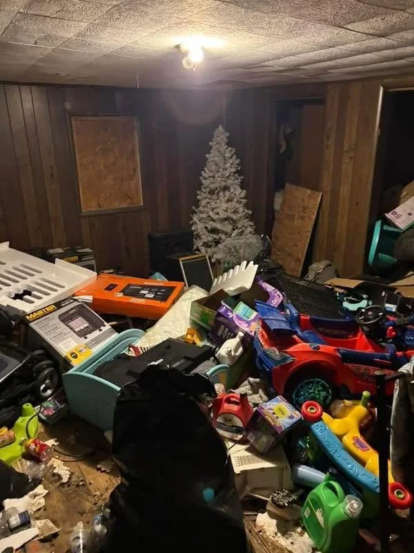A cluttered room with various toys and electronic items on a table, a white Christmas tree in the background, and wood-paneled walls.