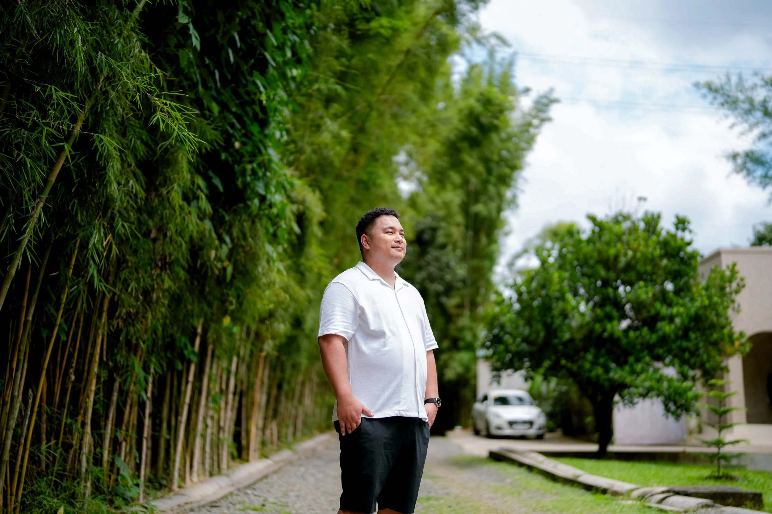 A man with short dark hair wearing a white polo shirt and black shorts standing outdoors on a sidewalk, with green trees and bushes on his left and parked cars and a building visible in the background.