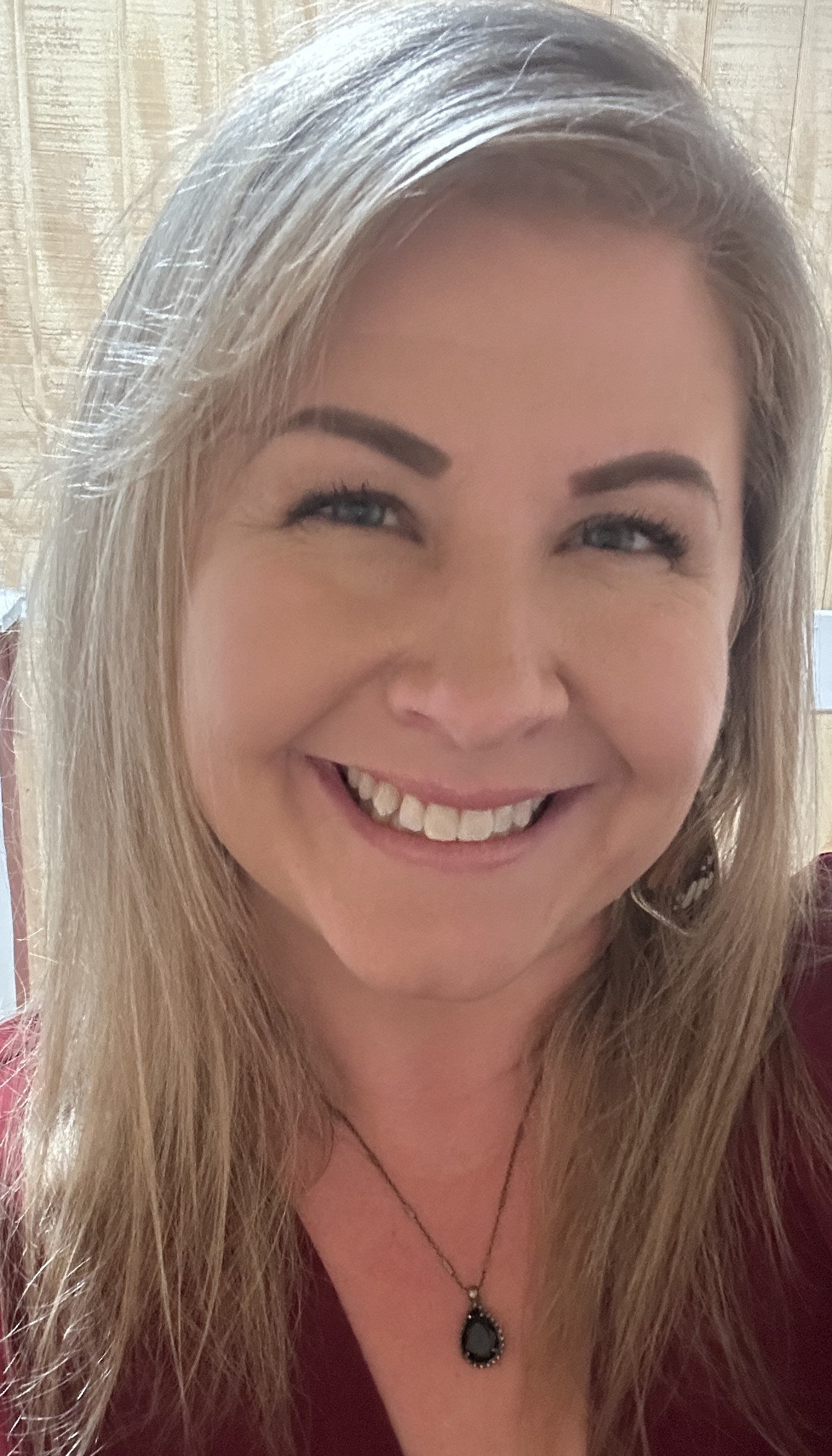 Close-up of a smiling woman with long blonde hair, wearing a dark necklace with a black pendant and a maroon top.
