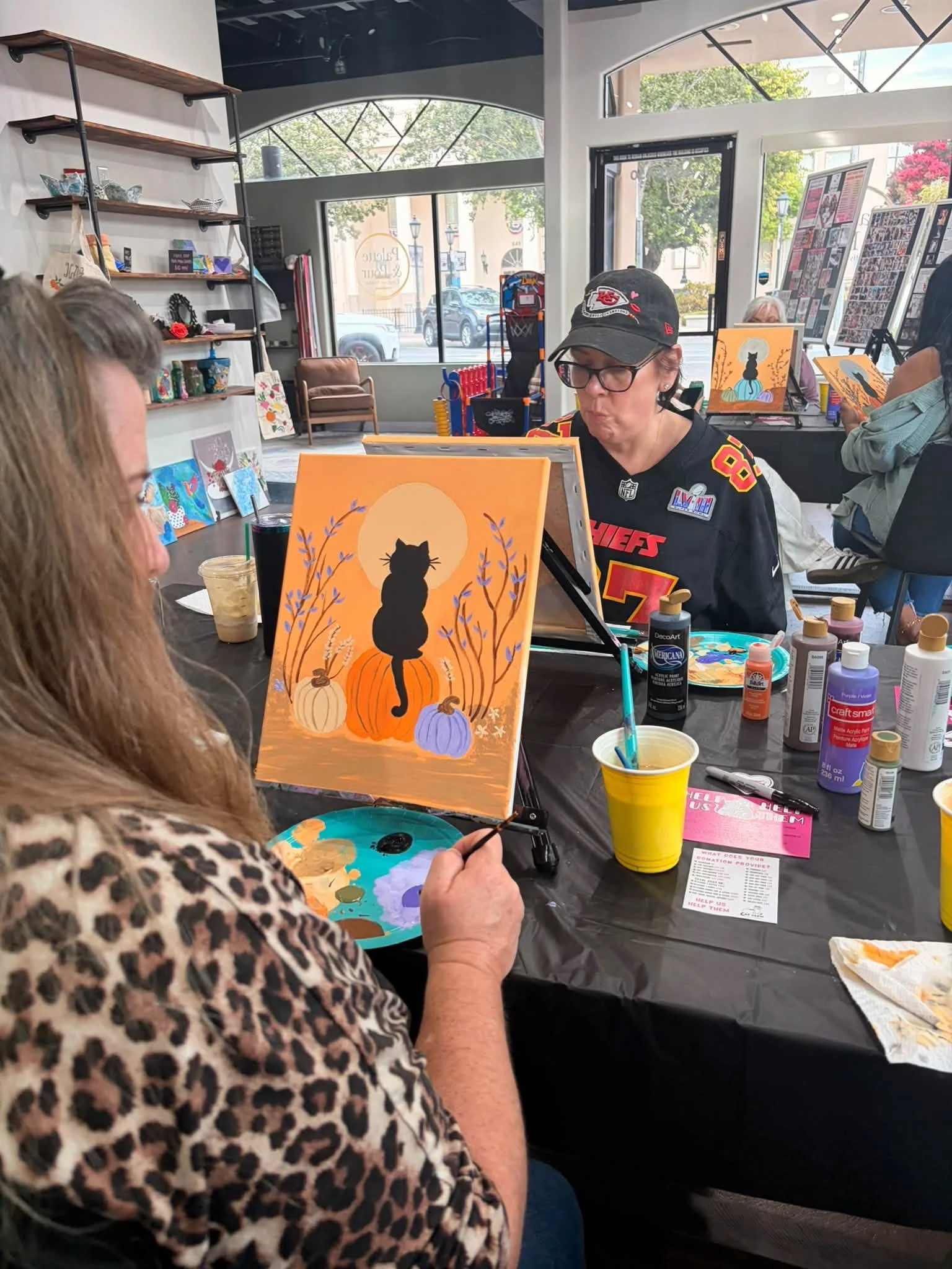 An indoor art class with people painting pictures of black cats sitting on pumpkins surrounded by fall leaves and pumpkins, with art supplies on the table.
