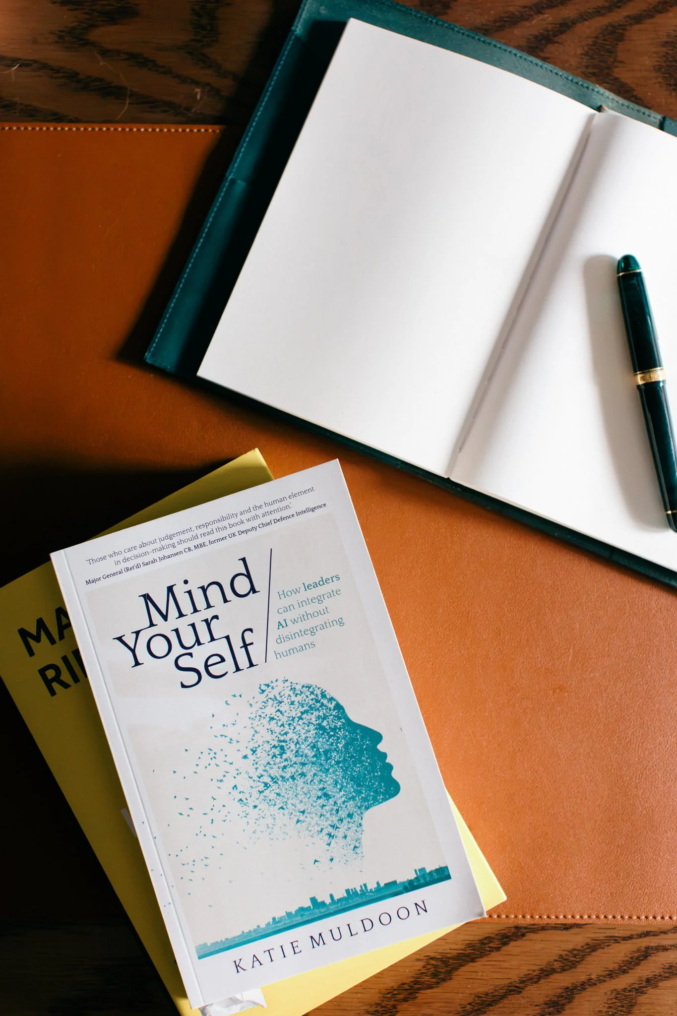 Open book titled 'Mind Your Own Mind' by Katie Muldoon next to a closed teal-colored notebook and a black pen, placed on a wooden surface.