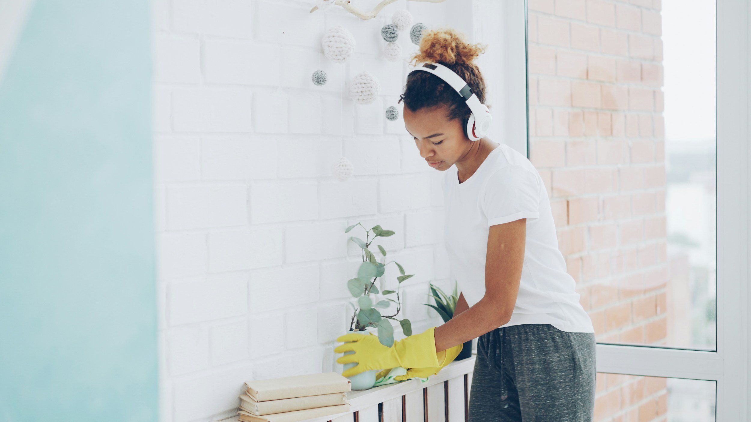 A woman wearing yellow rubber gloves and a white t-shirt is planting a small green plant in a pot on a white shelf, with a white wall and brick wall background.