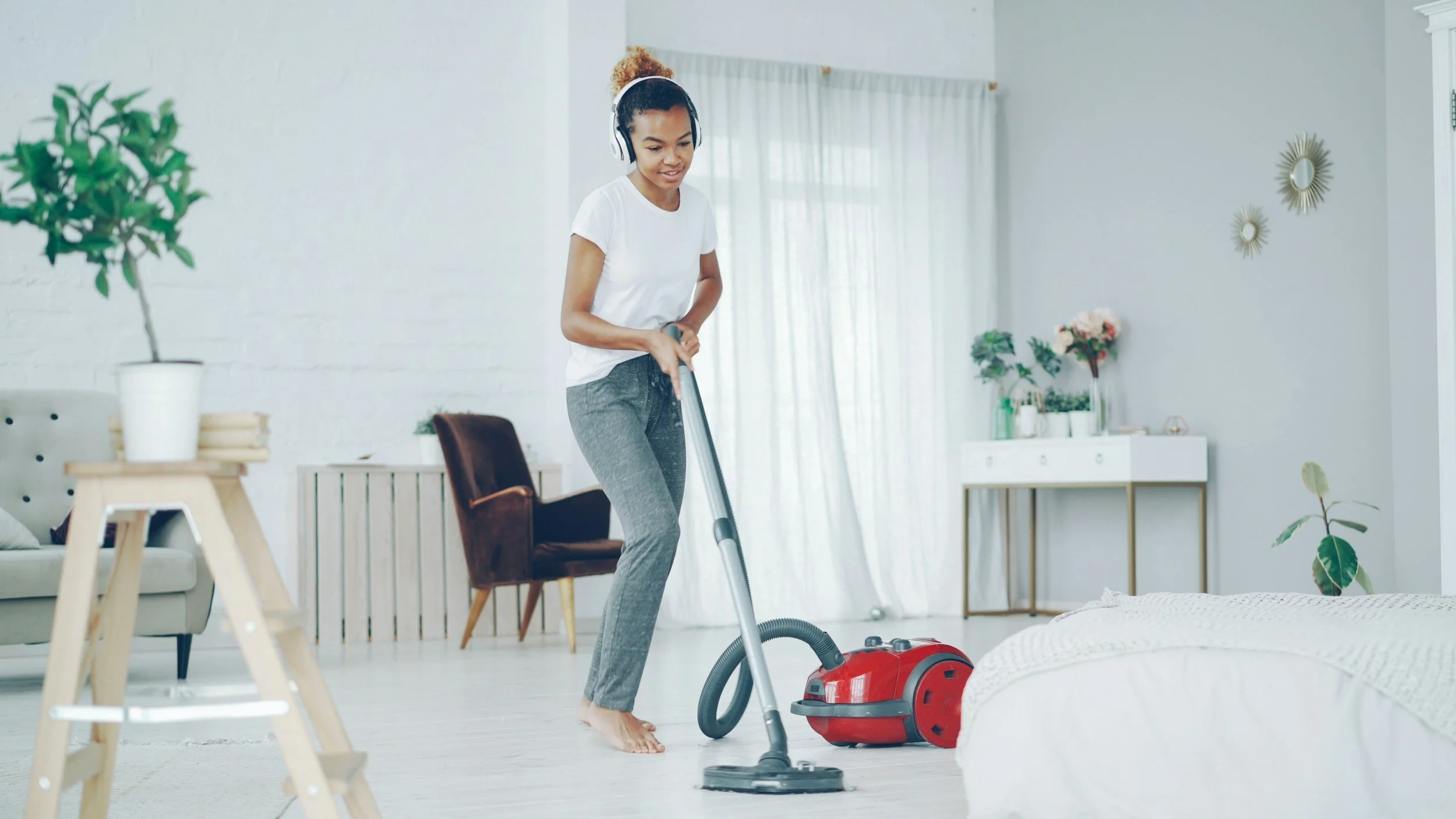 Young woman vacuuming in a bright, modern living room with plants and decorative wall mirrors.