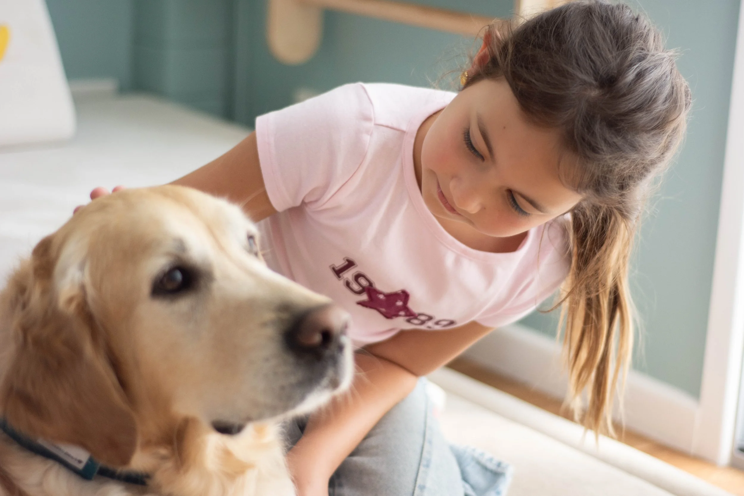 A girl with long brown hair wearing a pink T-shirt is cuddling a golden retriever puppy indoors. The girl is smiling and looking down at the puppy, which is looking slightly to the side.