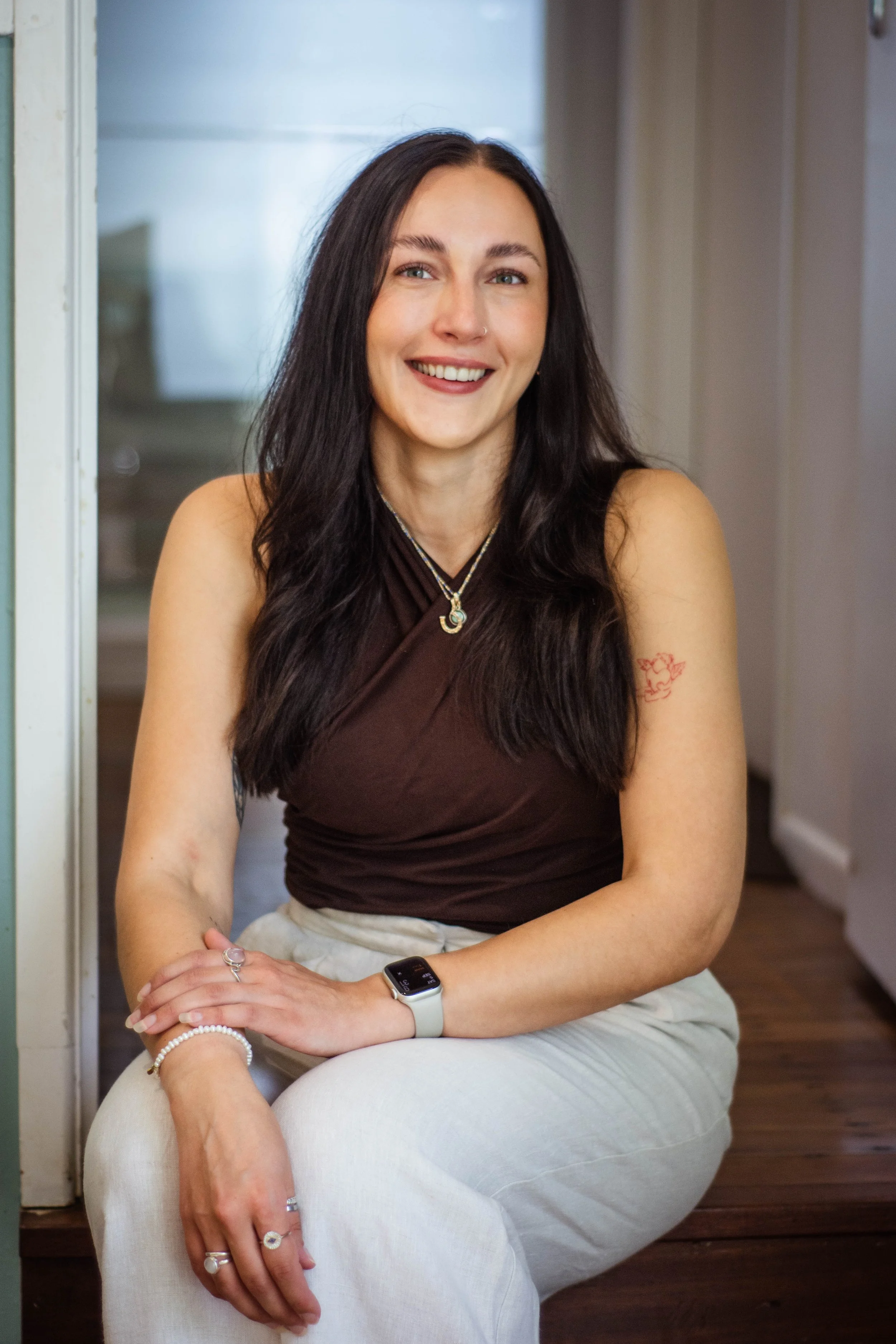 A young woman with long dark hair and a smile, sitting cross-legged on a wooden floor inside a house. She is wearing a dark brown sleeveless top, white pants, a silver necklace, an Apple Watch, and multiple rings. She has a small red tattoo on her right upper arm and a nose ring.