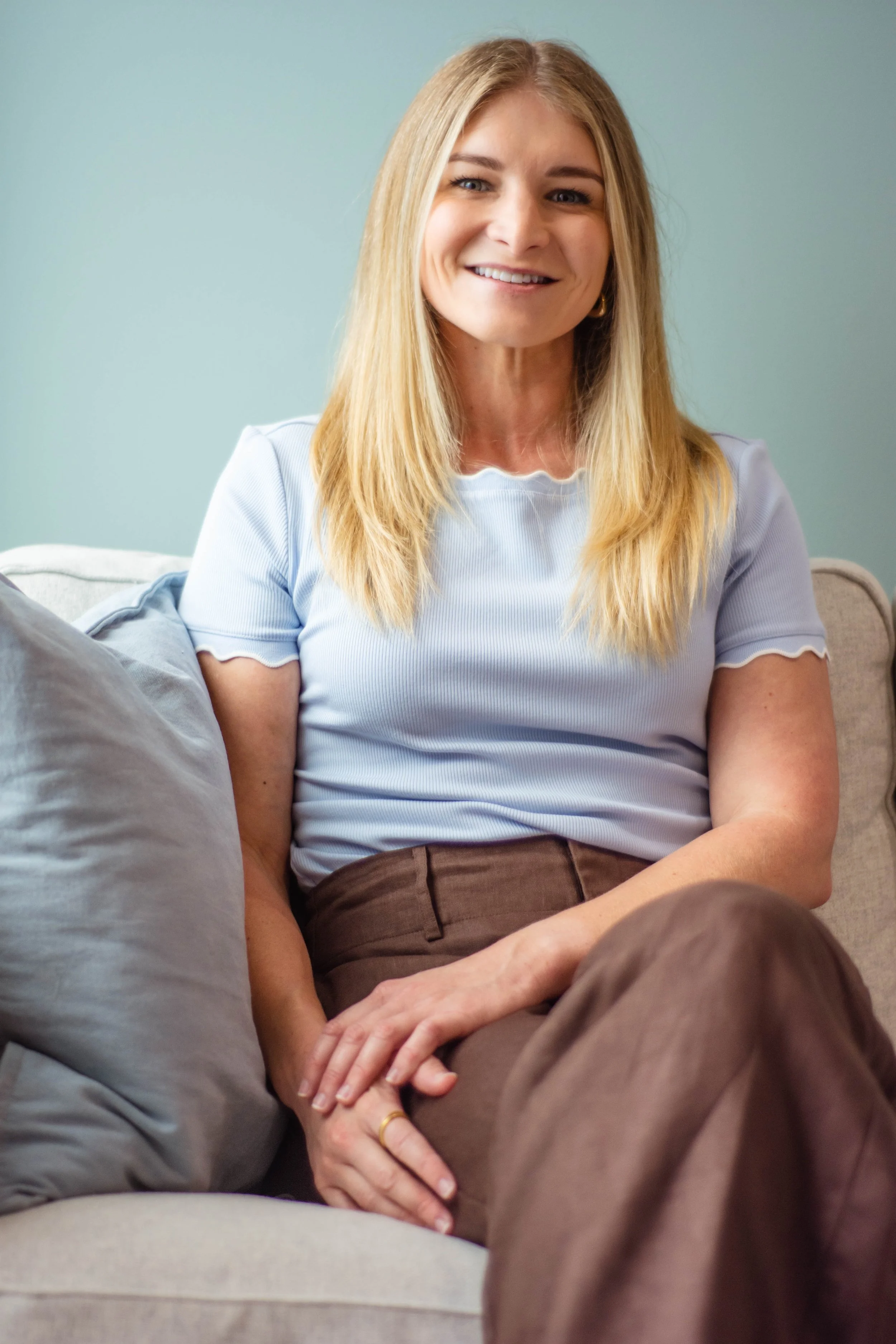 A woman with long blonde hair, smiling, sitting on a beige couch, wearing a light blue top and brown pants, in a room with light blue walls.