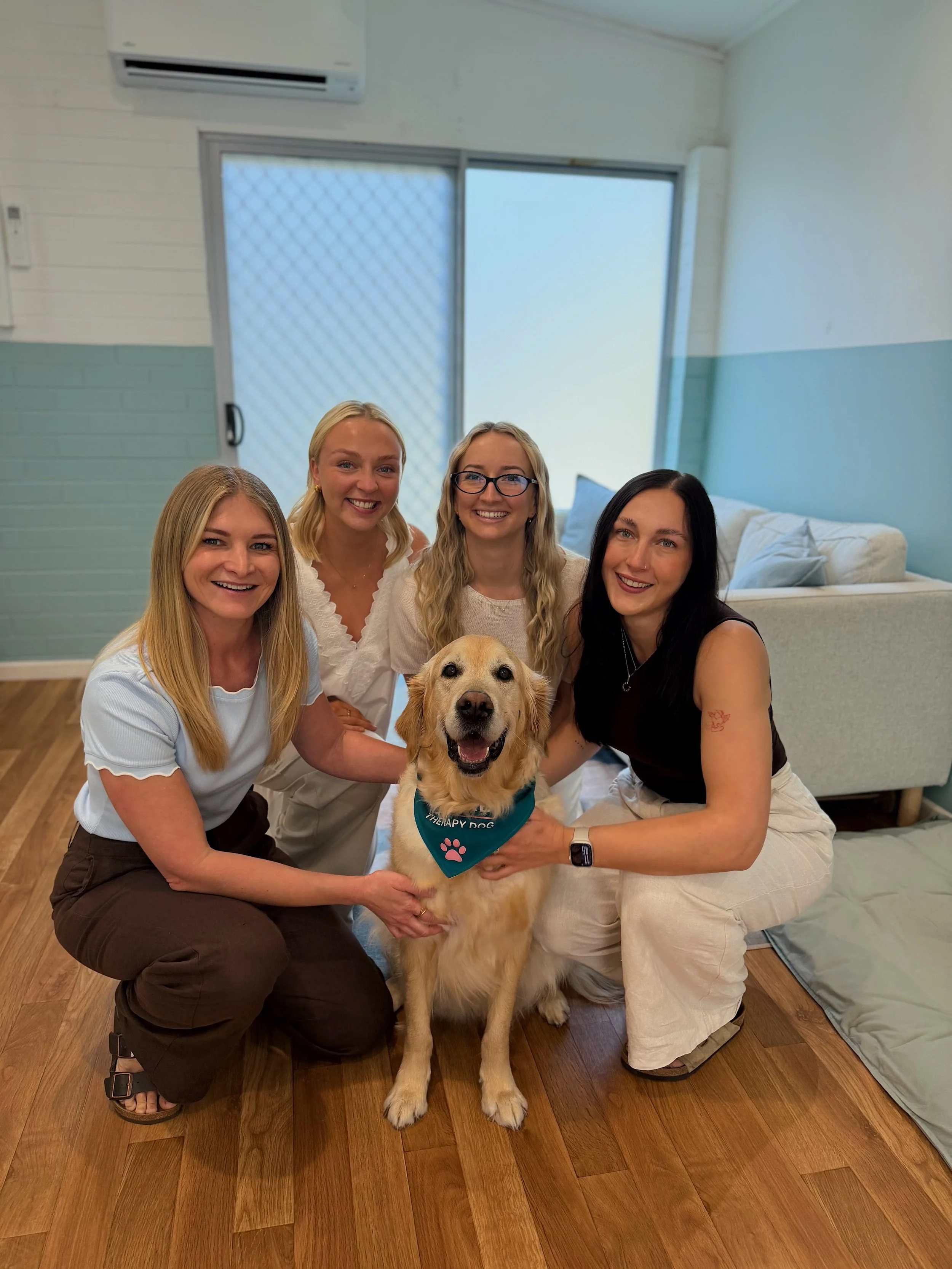 Four women and a service dog posing indoors, with a couch and window in the background.