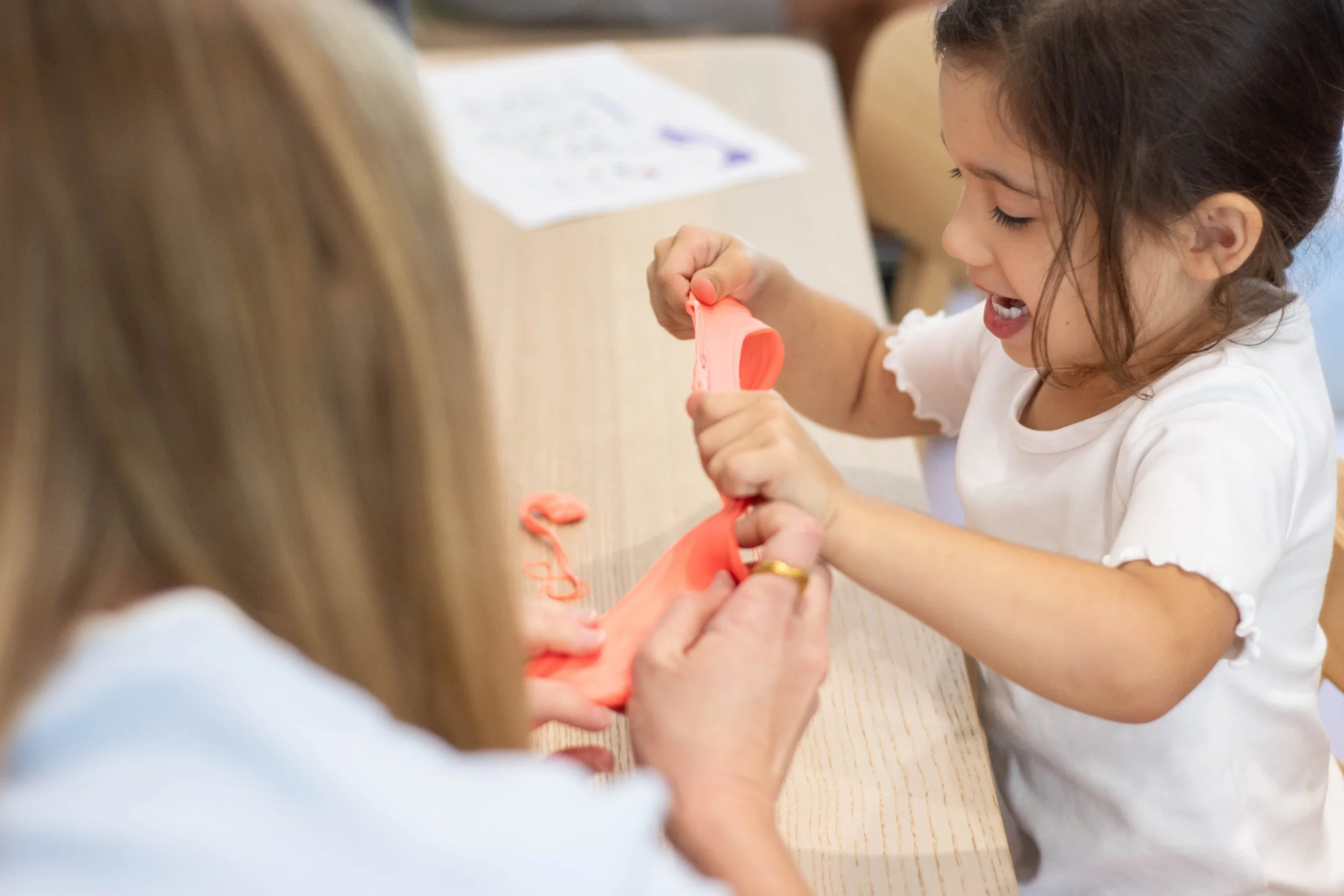 A young girl with dark hair and a white shirt plays with pink modeling clay at a table in a classroom or activity room.