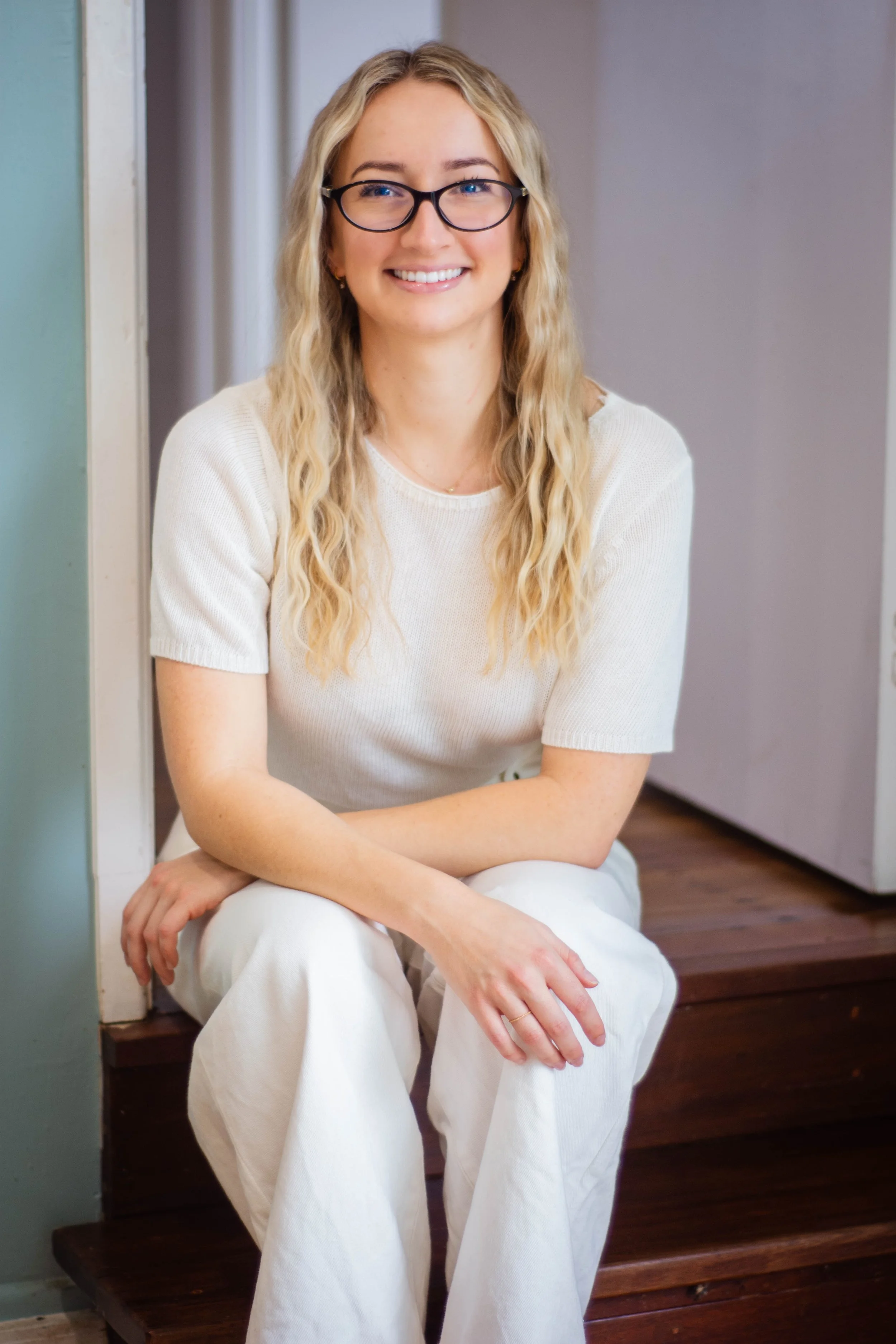 A young woman with blonde wavy hair, wearing glasses, a white short-sleeve shirt, and white pants, sitting on wooden stairs and smiling at the camera.