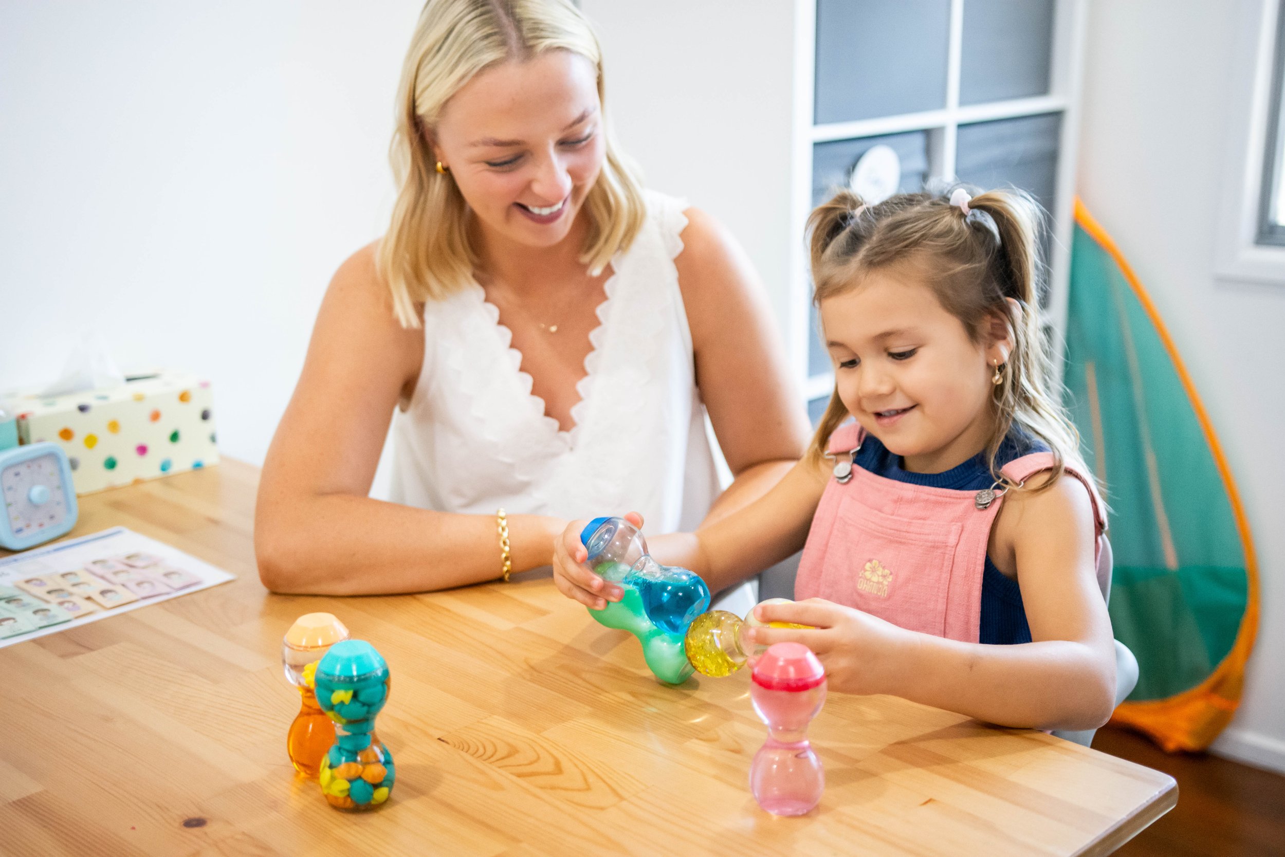 A woman and a young girl playing with colorful sand art bottles at a wooden table in a bright room.
