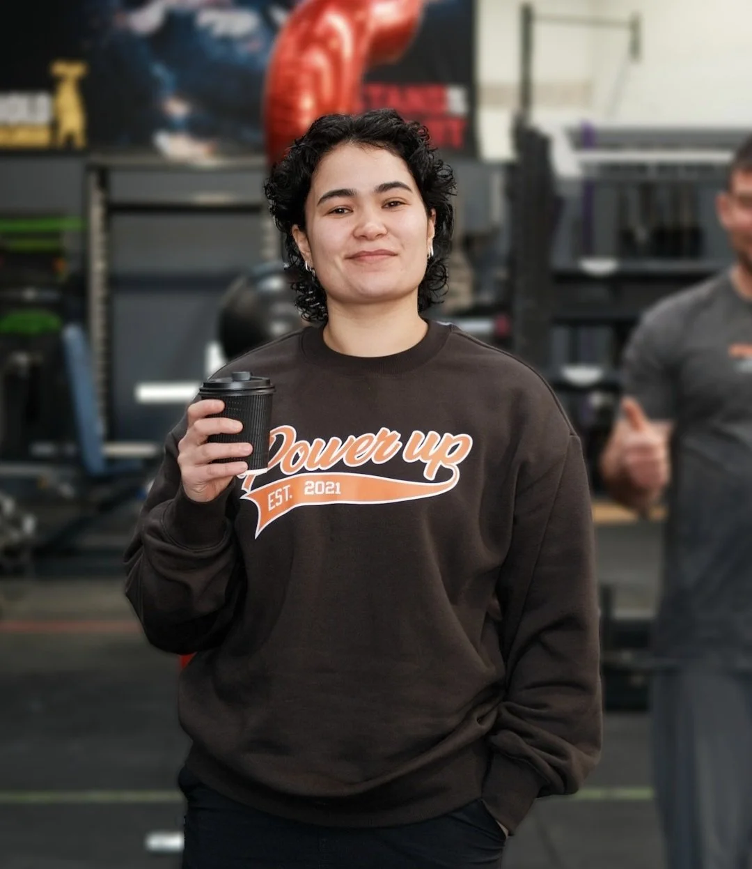 A young woman with short, curly black hair and earrings, smiling and holding a black coffee cup, standing in a gym with fitness equipment in the background, wearing a black sweatshirt with orange and white text that reads "Power up EST. 2021".