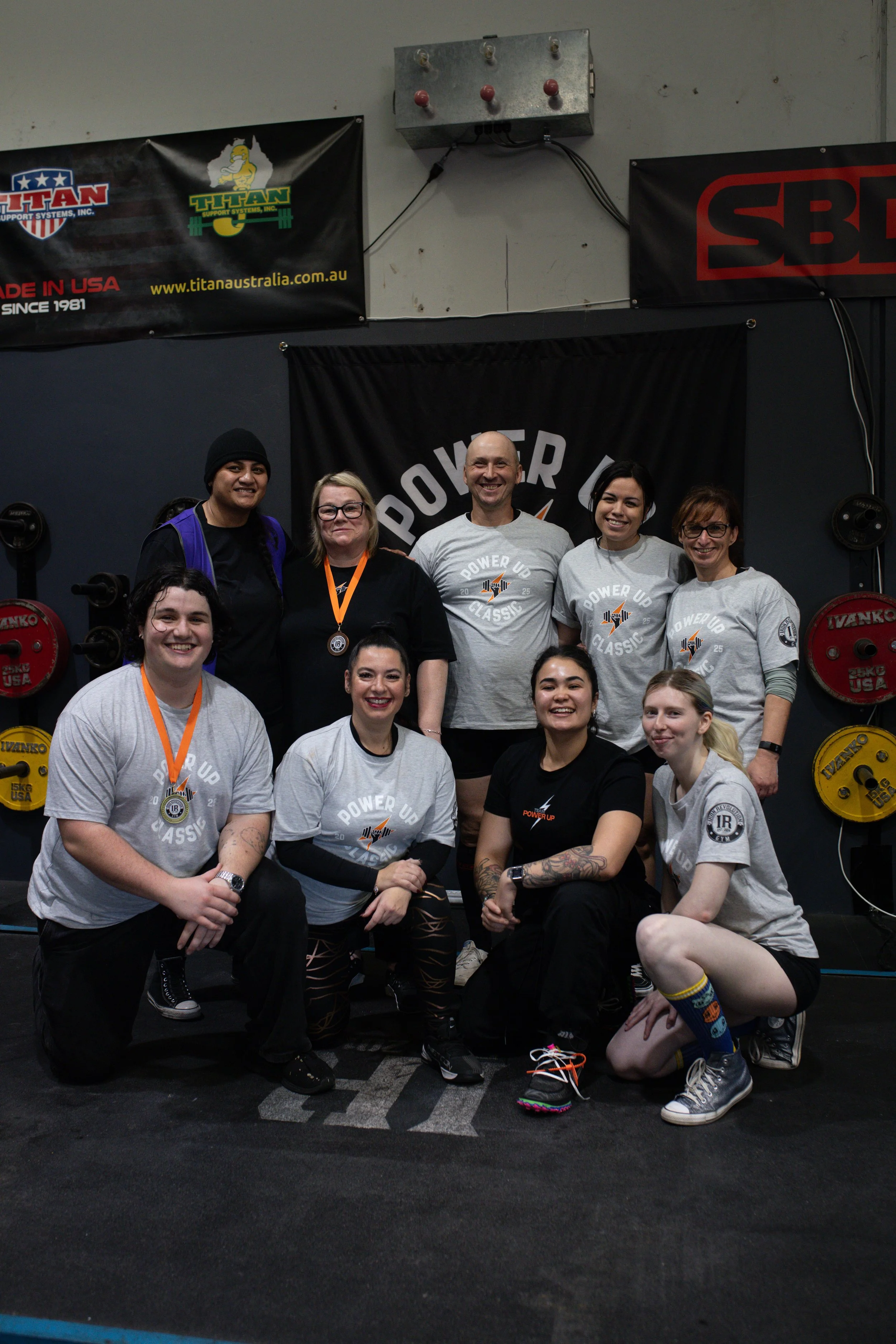 Group of people at a powerlifting event wearing T-shirts that say 'Power Up Classic,' with weightlifting equipment and banners in the background.