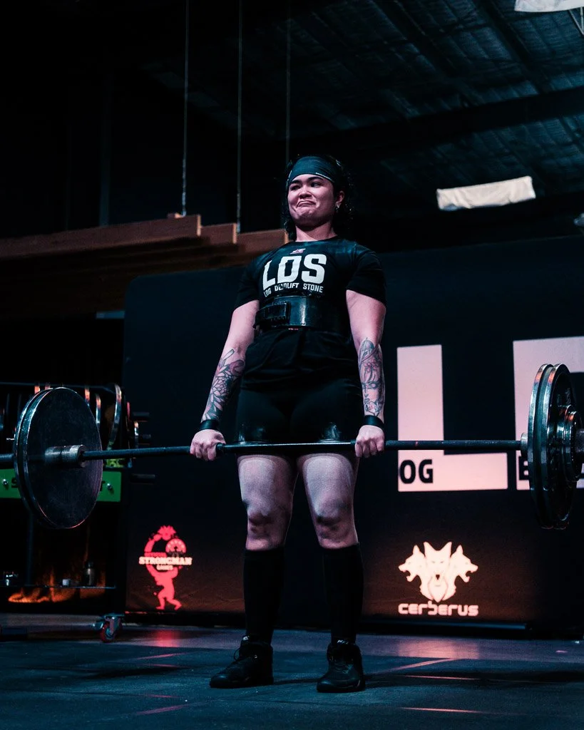 A female weightlifter with tattoos on her arms is lifting a barbell in a gym or competition setting. She is wearing a black shirt, shorts, socks, and a headband, and appears focused.