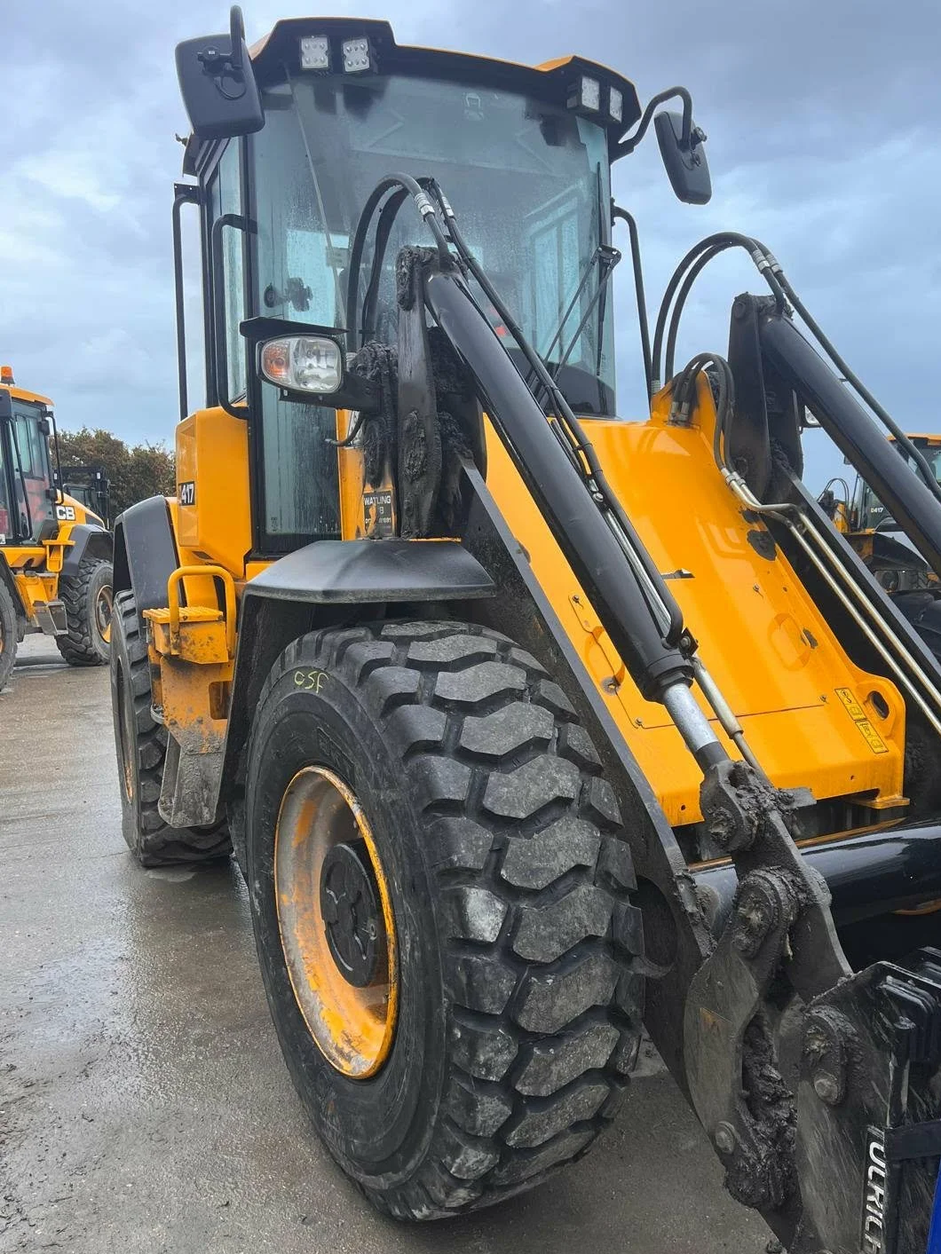 A yellow front loader construction vehicle with large tires and hydraulic arms parked outdoors, possibly on a wet surface with cloudy sky.