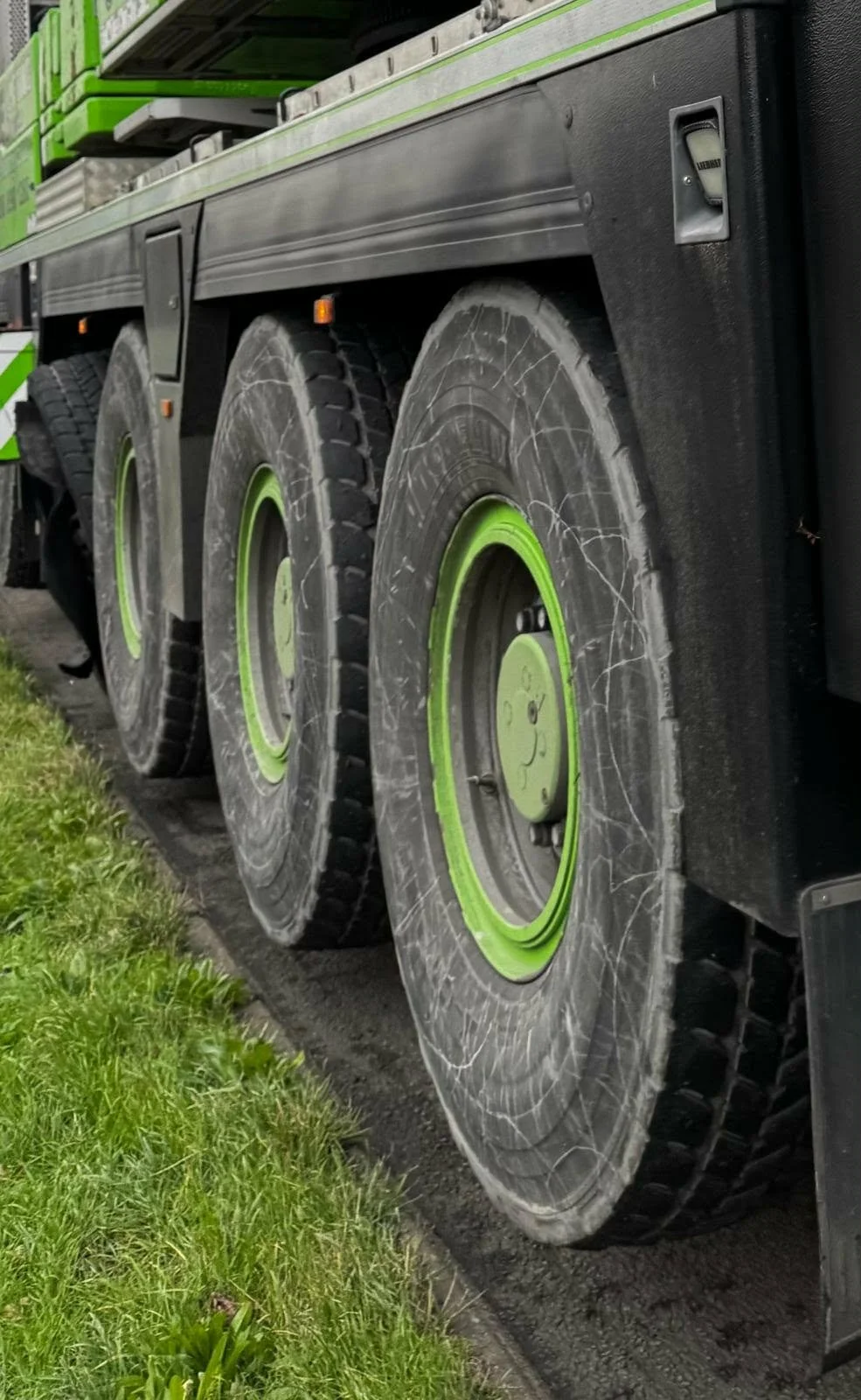 Close-up of large truck wheels with green accents, showing tire scratches and mud, parked on the side of a grassy area.