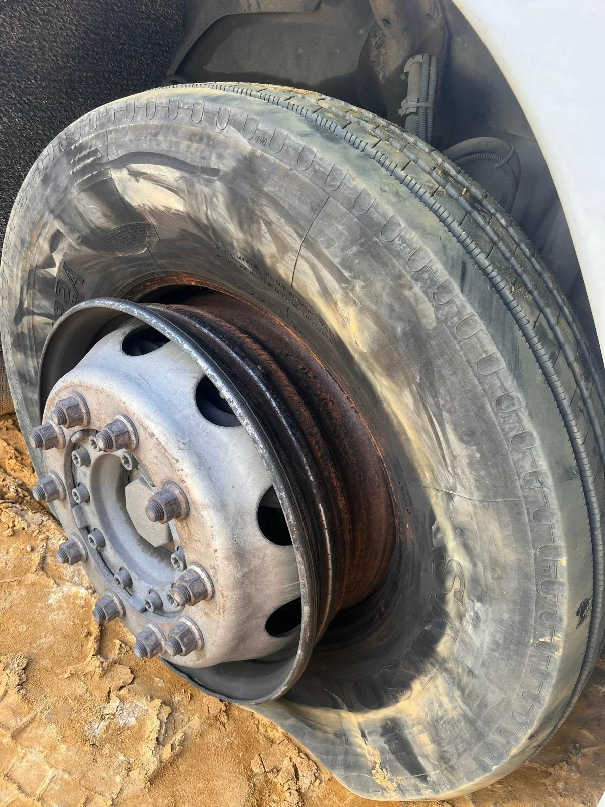 A close-up of a vehicle wheel with a tire mounted on a metallic rim, lying on sandy ground. The wheel shows signs of rust and wear.