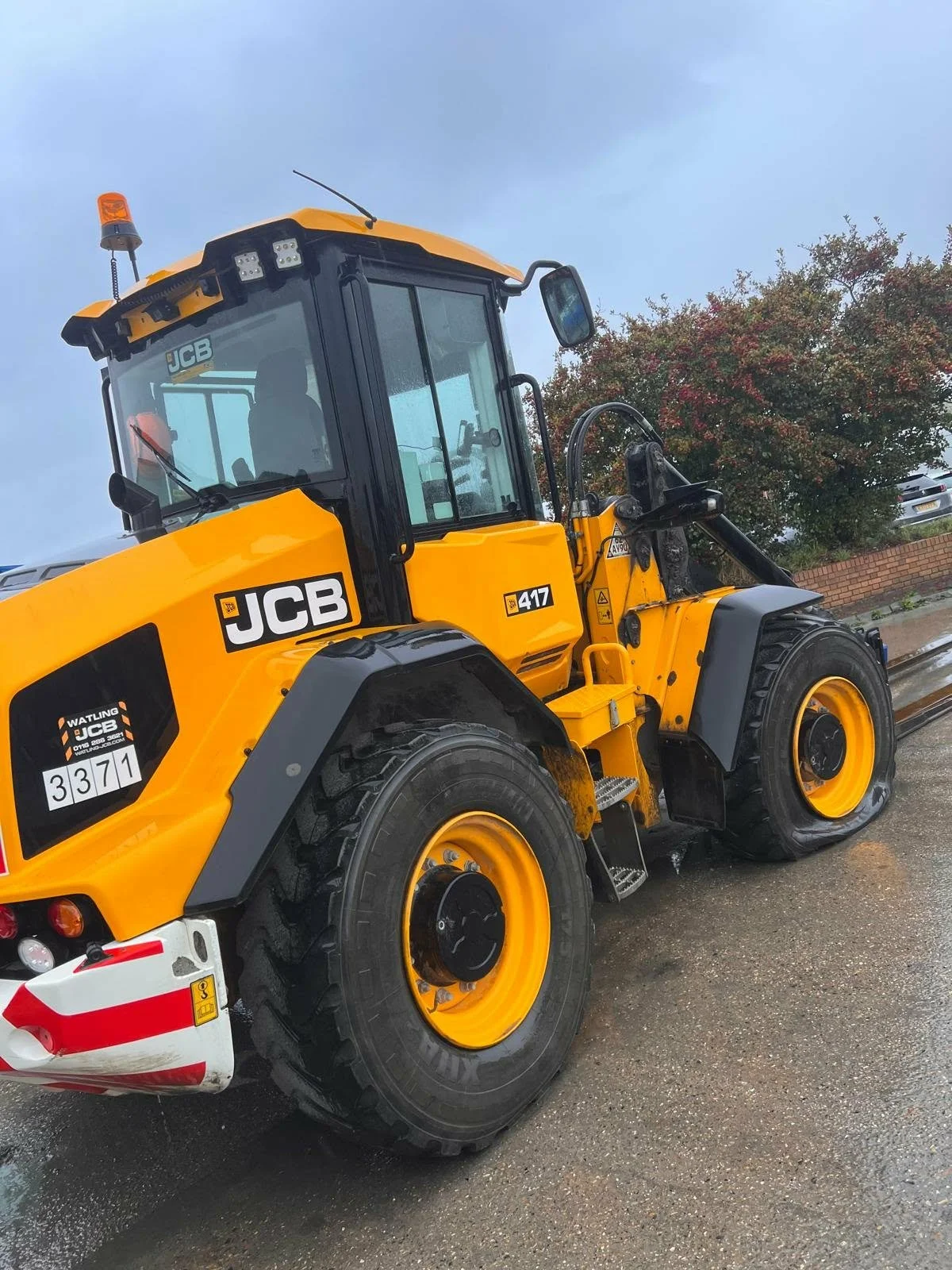 A yellow JCB model 417 wheel loader parked on a wet pavement with a brick wall and trees in the background.