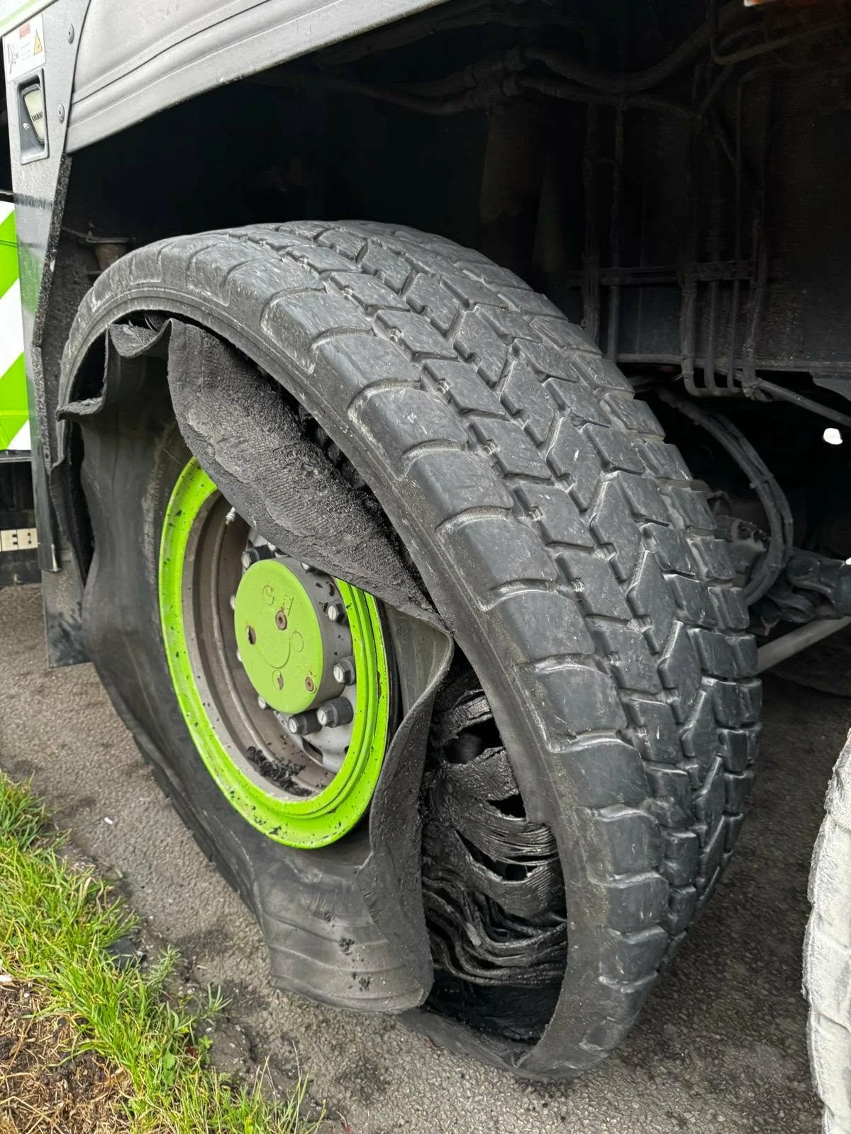 Image of a large truck tire with a damaged and deflated tire sidewall, exposing the internal structure, next to a patch of grass on a paved surface.