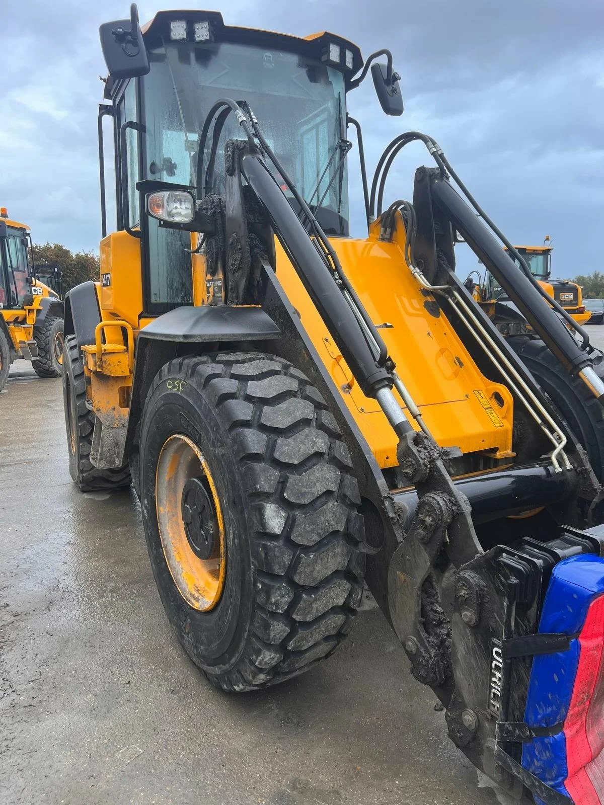 A yellow and black construction backhoe loader with a large front wheel, parked outdoors on a wet surface. Several similar vehicles are in the background.