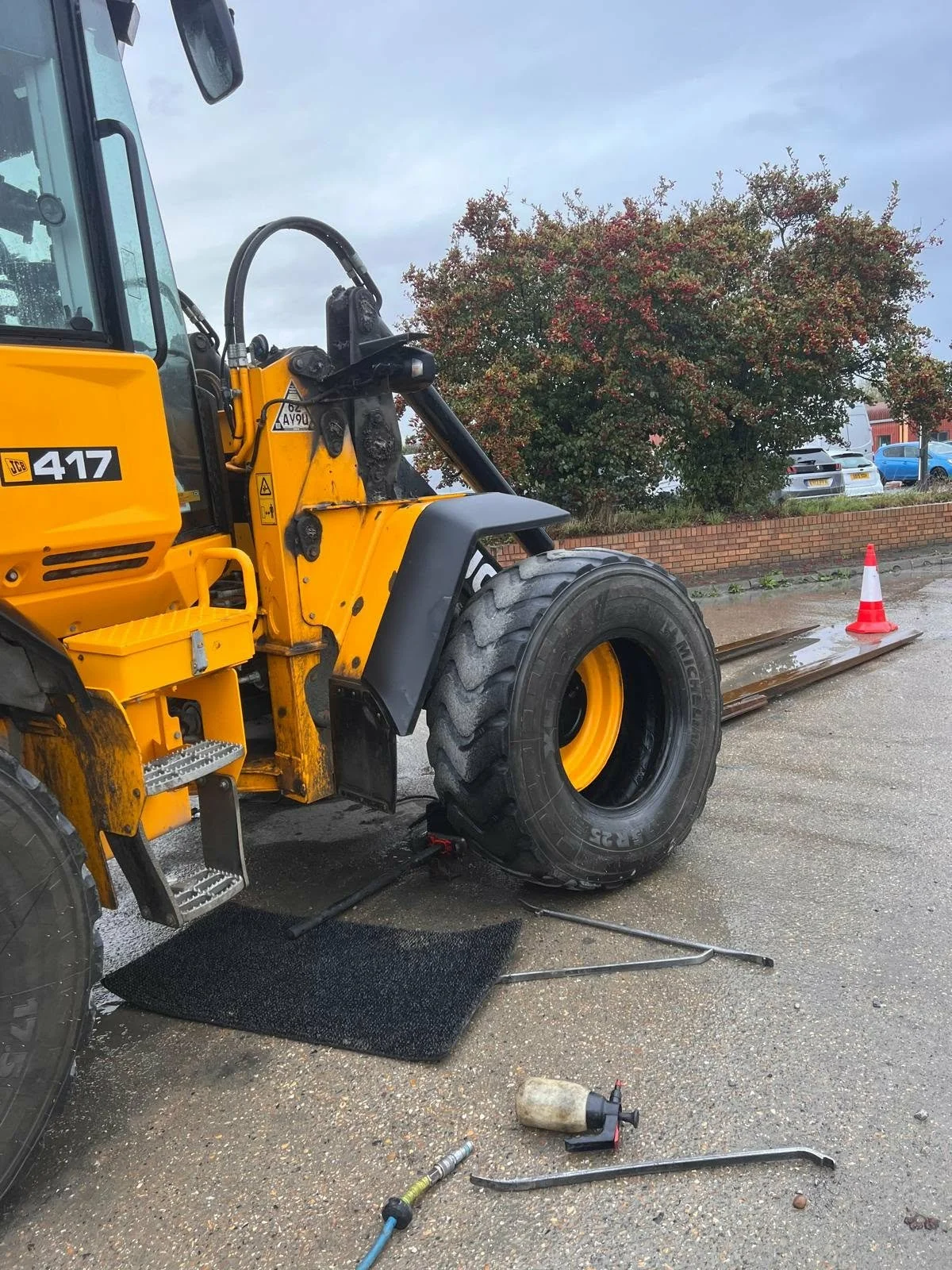 A yellow JCB 417 wheel loader parked on a wet surface with maintenance tools around, including a hydraulic jack, a tire iron, and a fluid container. A traffic cone is placed nearby, and a tree with autumn foliage and a parking lot with cars are visib