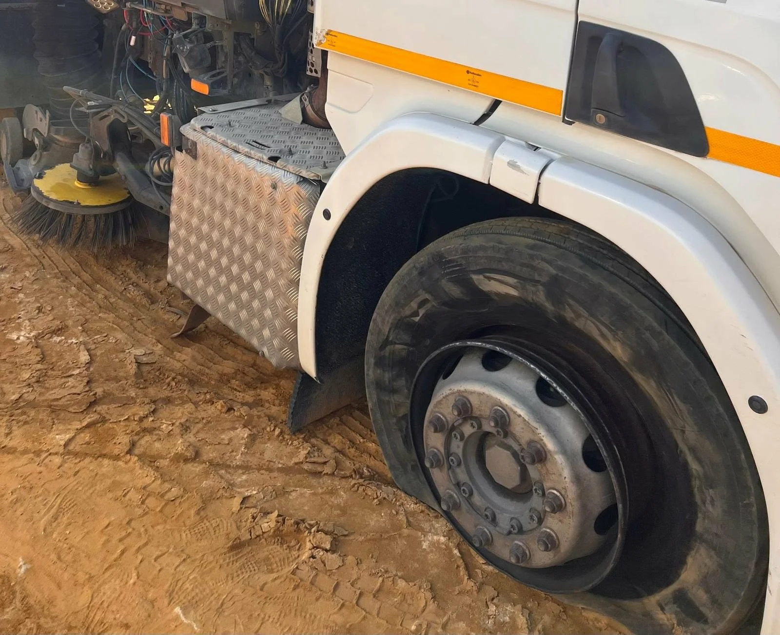 Close-up of the front part of a white street maintenance truck with a large black tire and a brush attachment on a dirt surface.