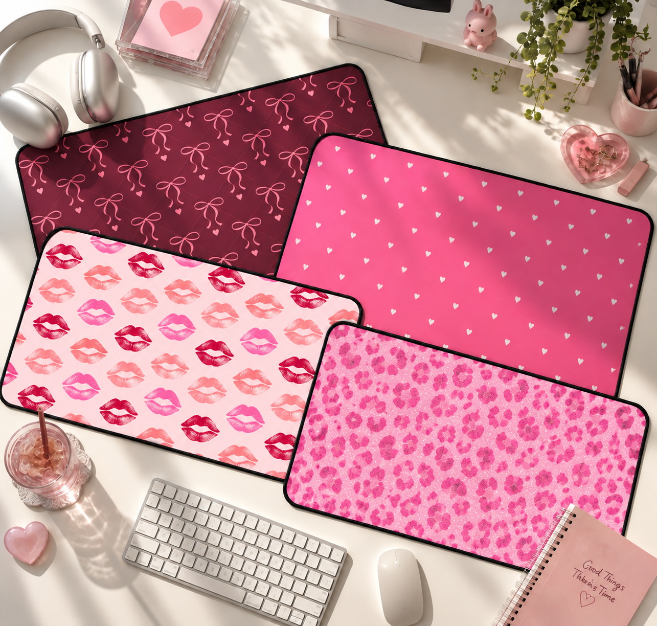 Desk setup with four pink-themed mouse pads with different patterns: bows, hearts, lips, and leopard print. Also includes a white keyboard, a white mouse, a glass of iced beverage with a pink straw, a closed pink notebook with writing, a small pink heart-shaped item, headphones, and decorative items like a small pink bunny figurine, green plants, and pink containers with pens and jewelry.