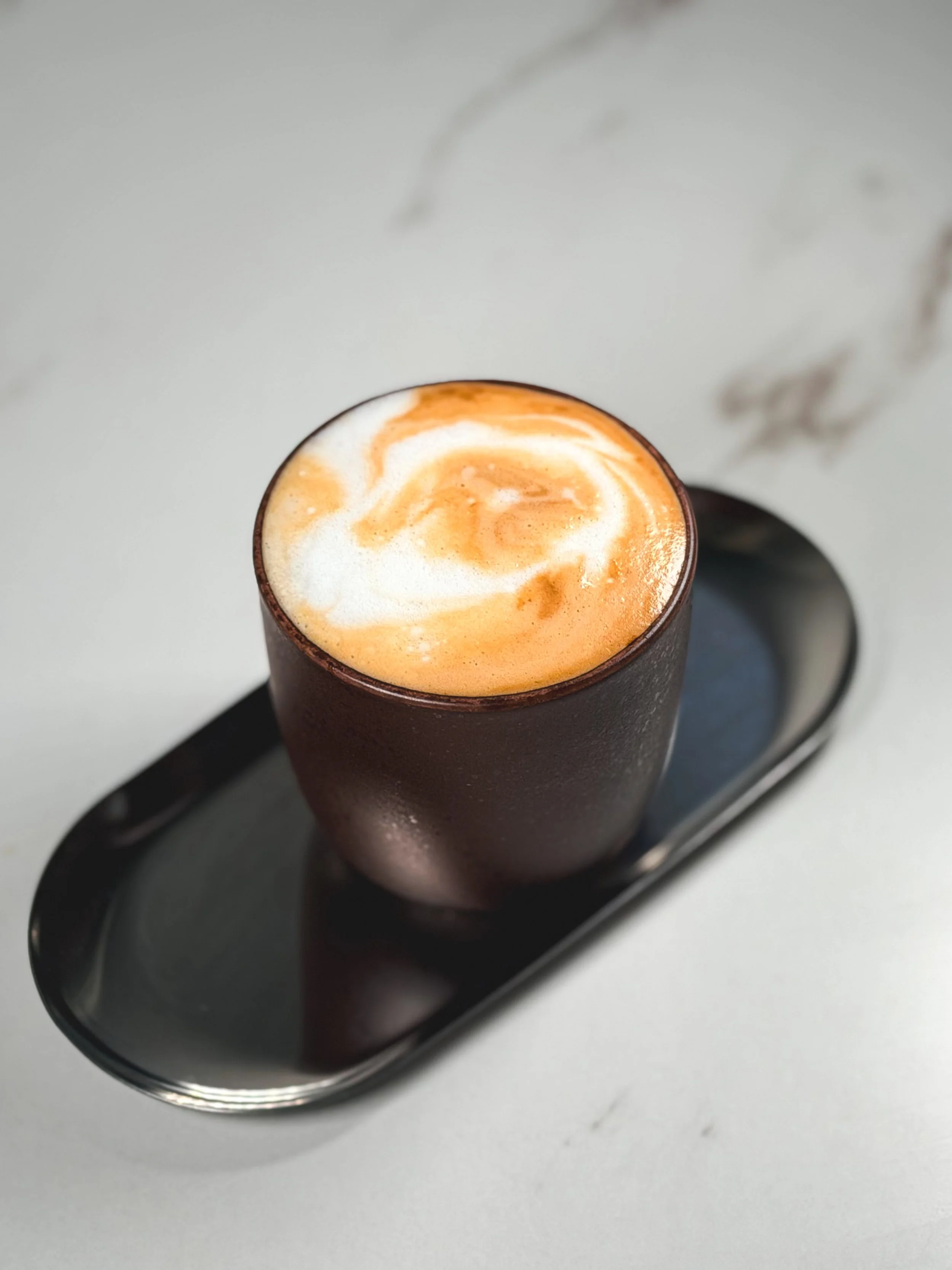 A black mug filled with a frothy coffee beverage, placed on a black tray with a spoon on the side, on a white surface.