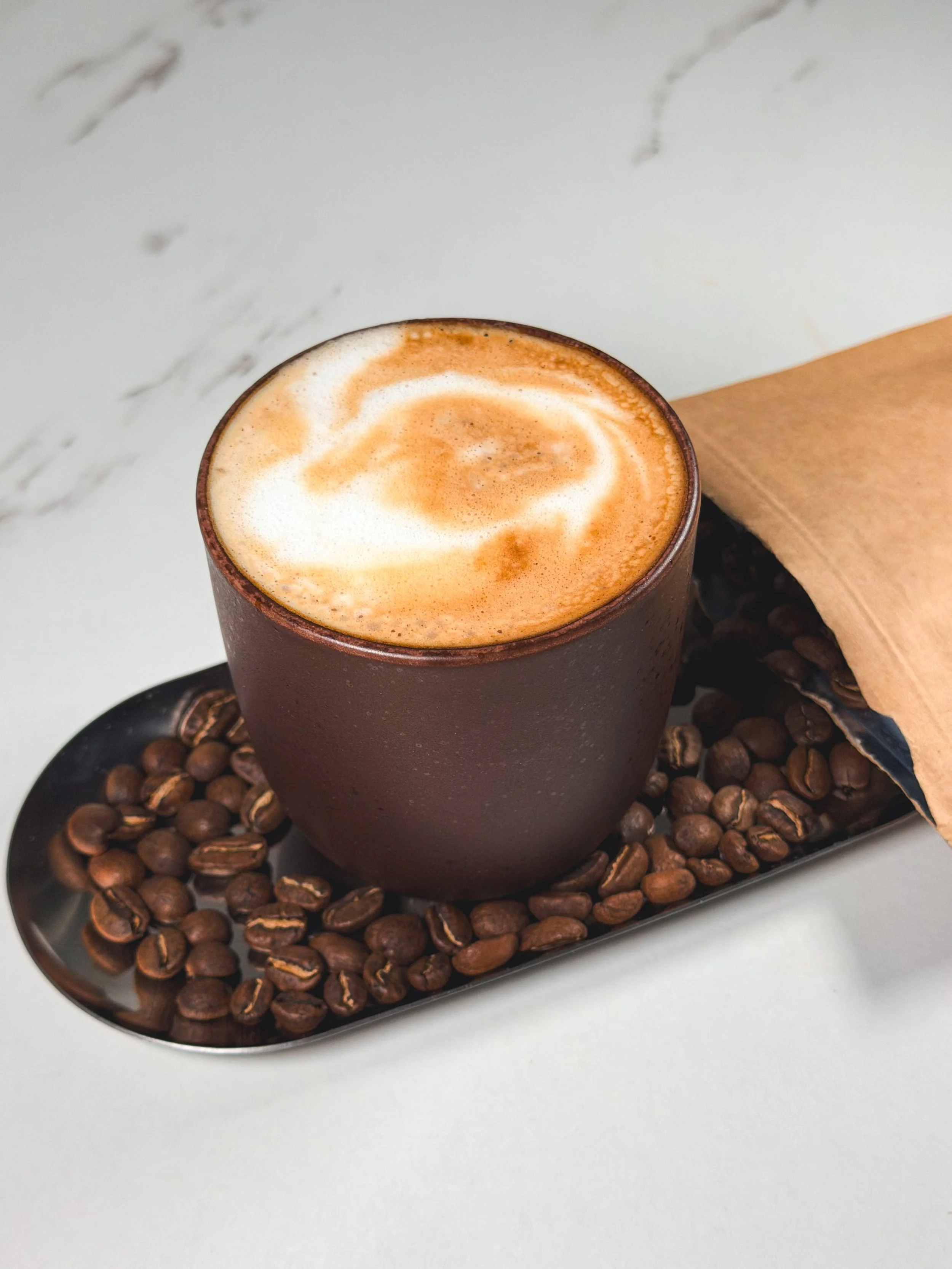 A brown ceramic cup filled with coffee topped with foam and cinnamon, on a black tray surrounded by coffee beans, with a piece of paper nearby.