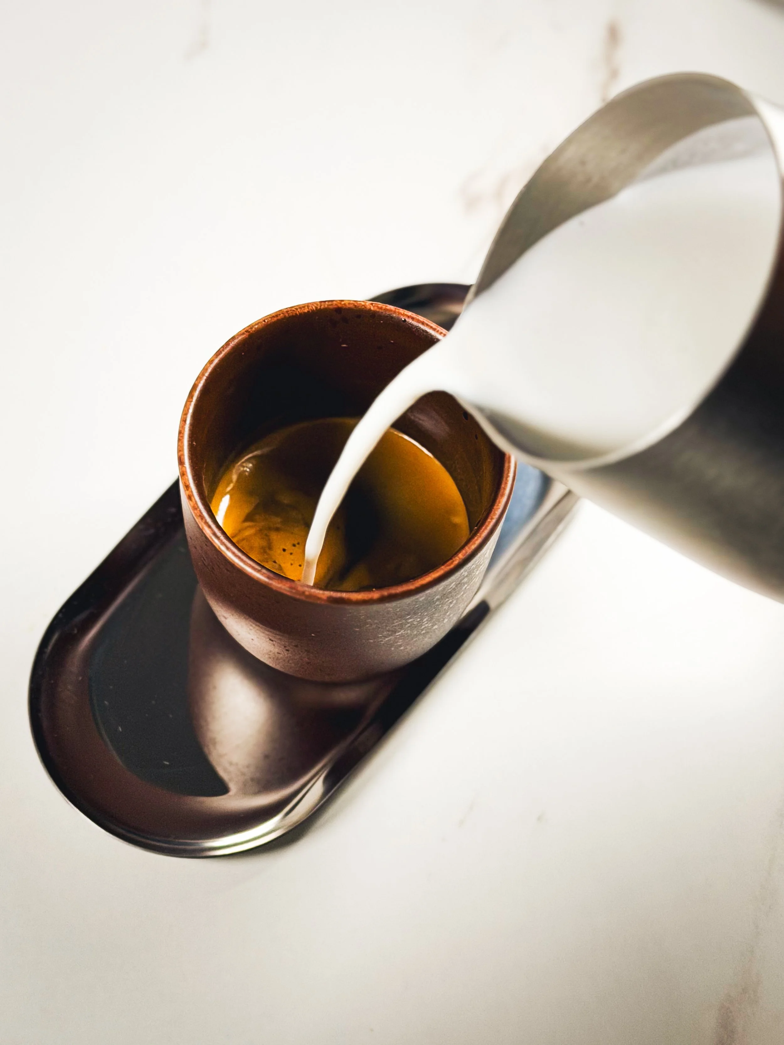 Pouring milk into a brown ceramic mug filled with black coffee on a black tray.