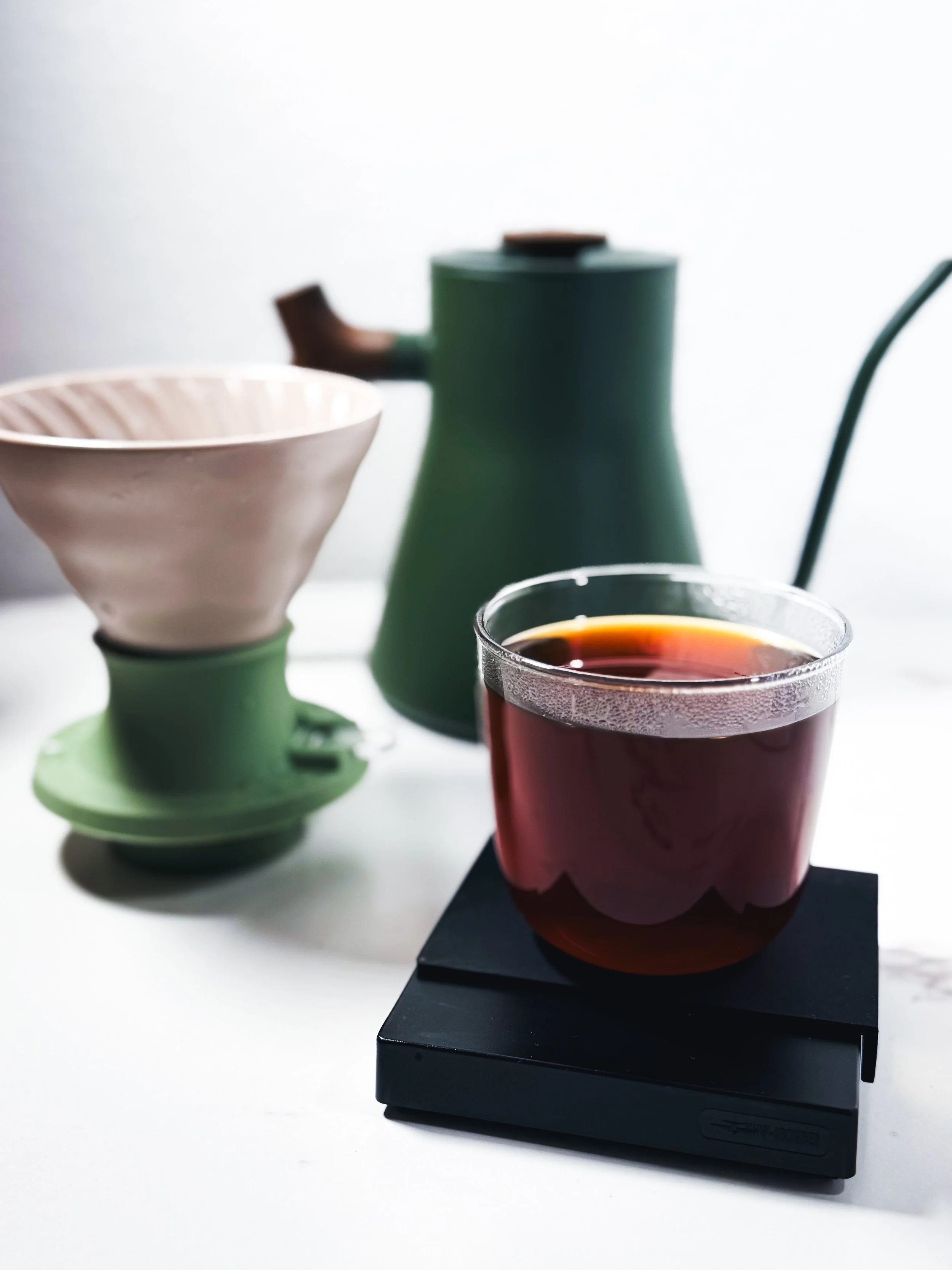 Close-up of a glass of tea and ceramic teapot on a black tray, with a pink bowl and a green teapot in the background.