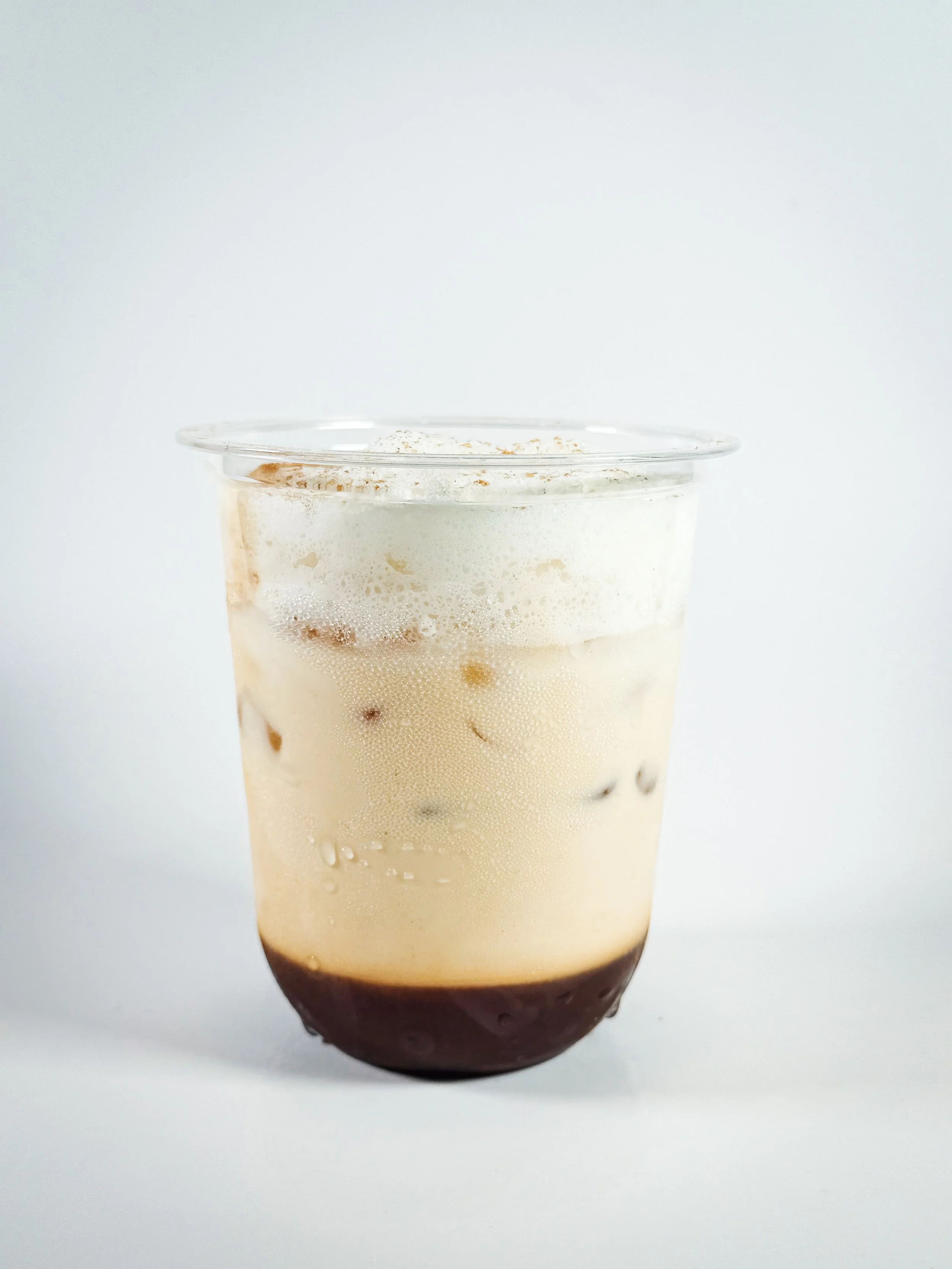 A glass of iced coffee with milk, topped with foam, and a layer of chocolate at the bottom against a plain white background.