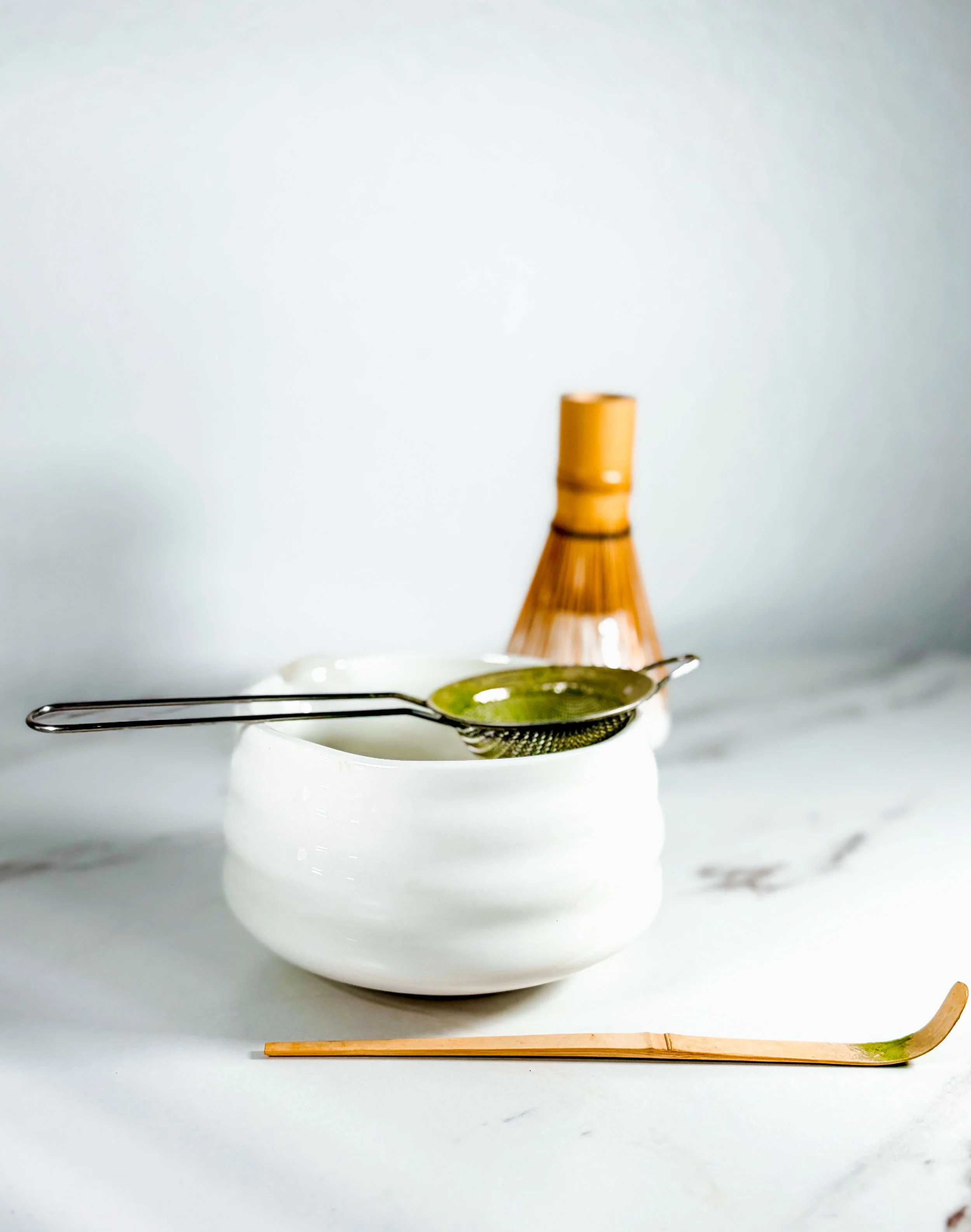 A ceramic bowl with a metal tea strainer resting on top, a pair of wooden chopsticks lying on the white surface, and a small portion of a brown bottle with a golden cap visible in the background.